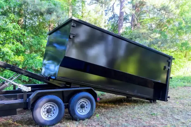 Black dump trailer with dual axles parked on a wooded slope outdoors
