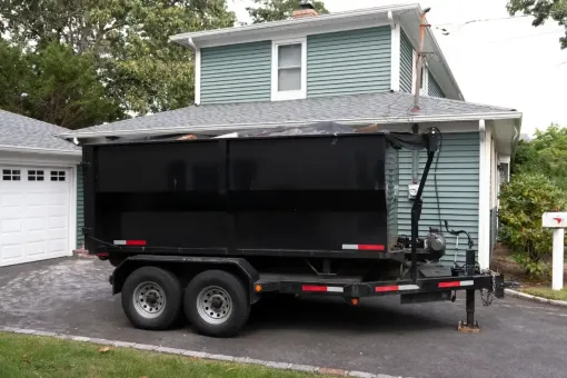 Black dump trailer parked in a driveway beside a gray house and garage.