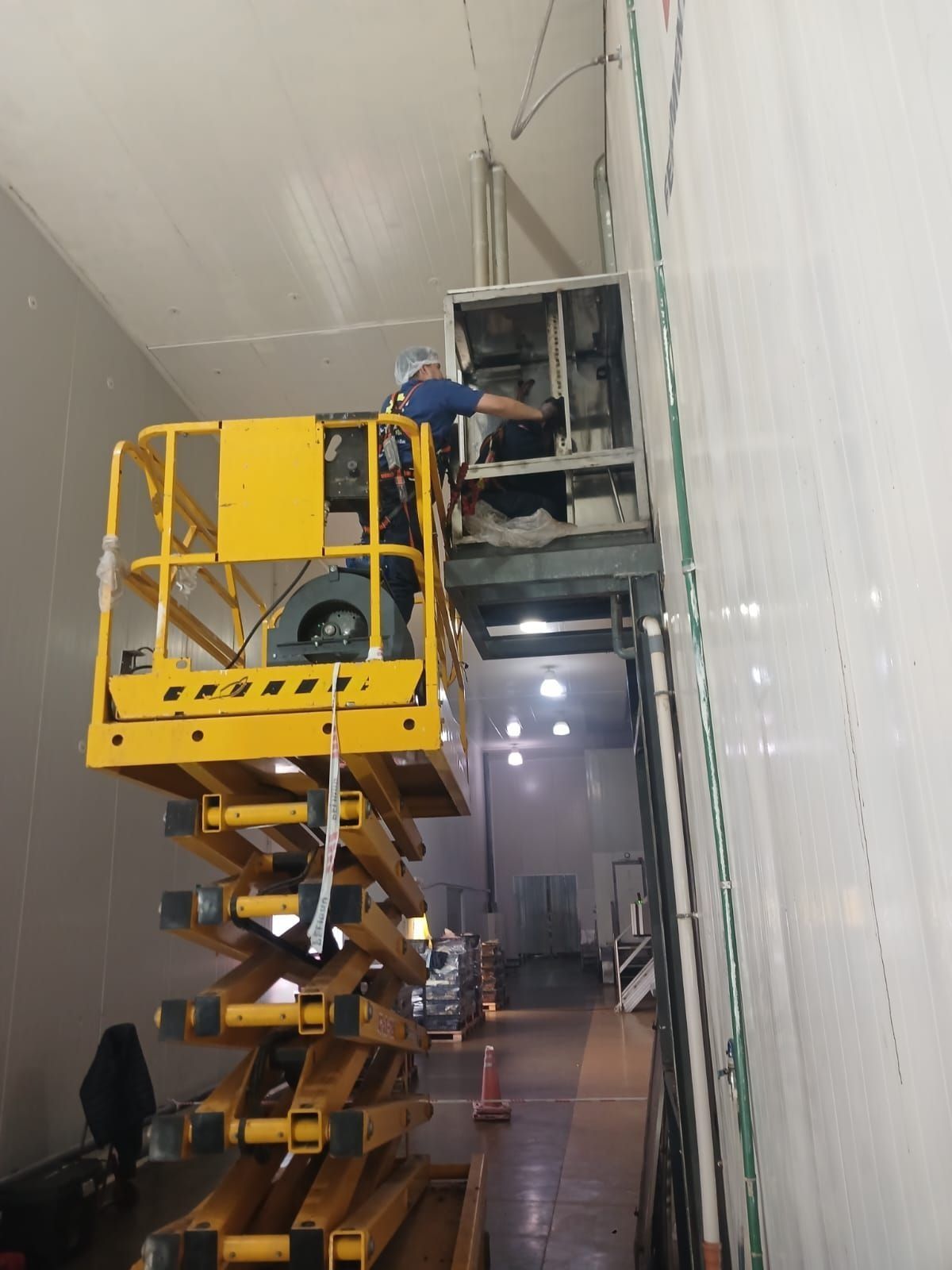 Un operario repara equipos en una plataforma elevadora de tijera amarilla en un estrecho pasillo industrial.