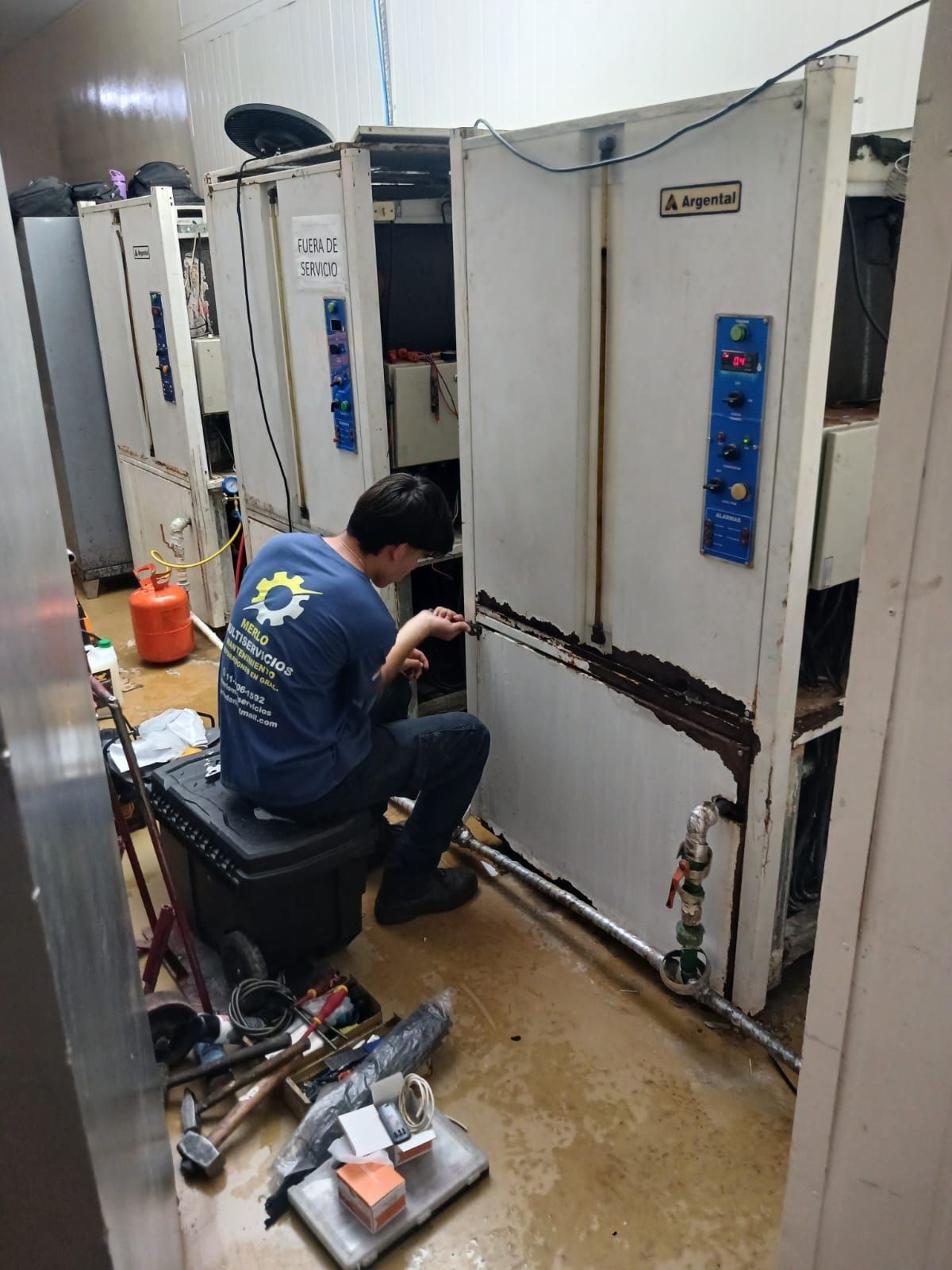 Un técnico se arrodilla junto a una gran máquina industrial en un taller húmedo, inspeccionando su panel inferior.