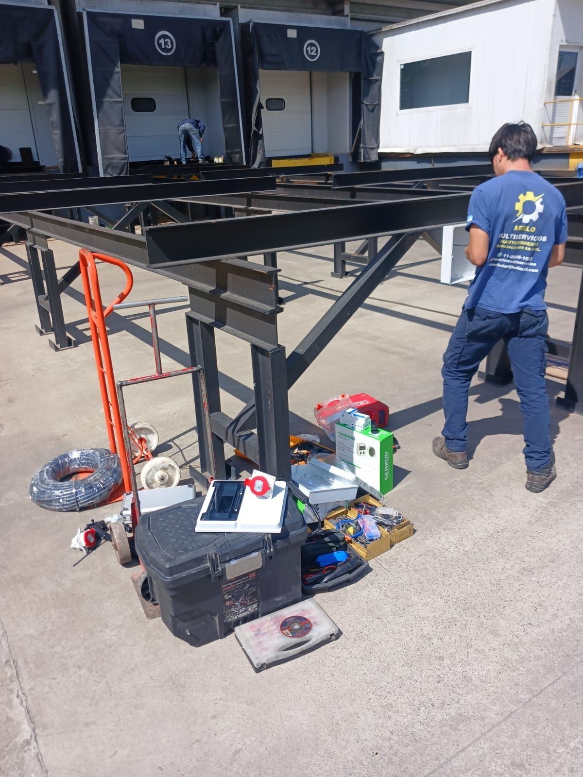Un hombre trabaja junto a la estructura metálica negra de un remolque, con herramientas en el suelo, en un patio industrial.