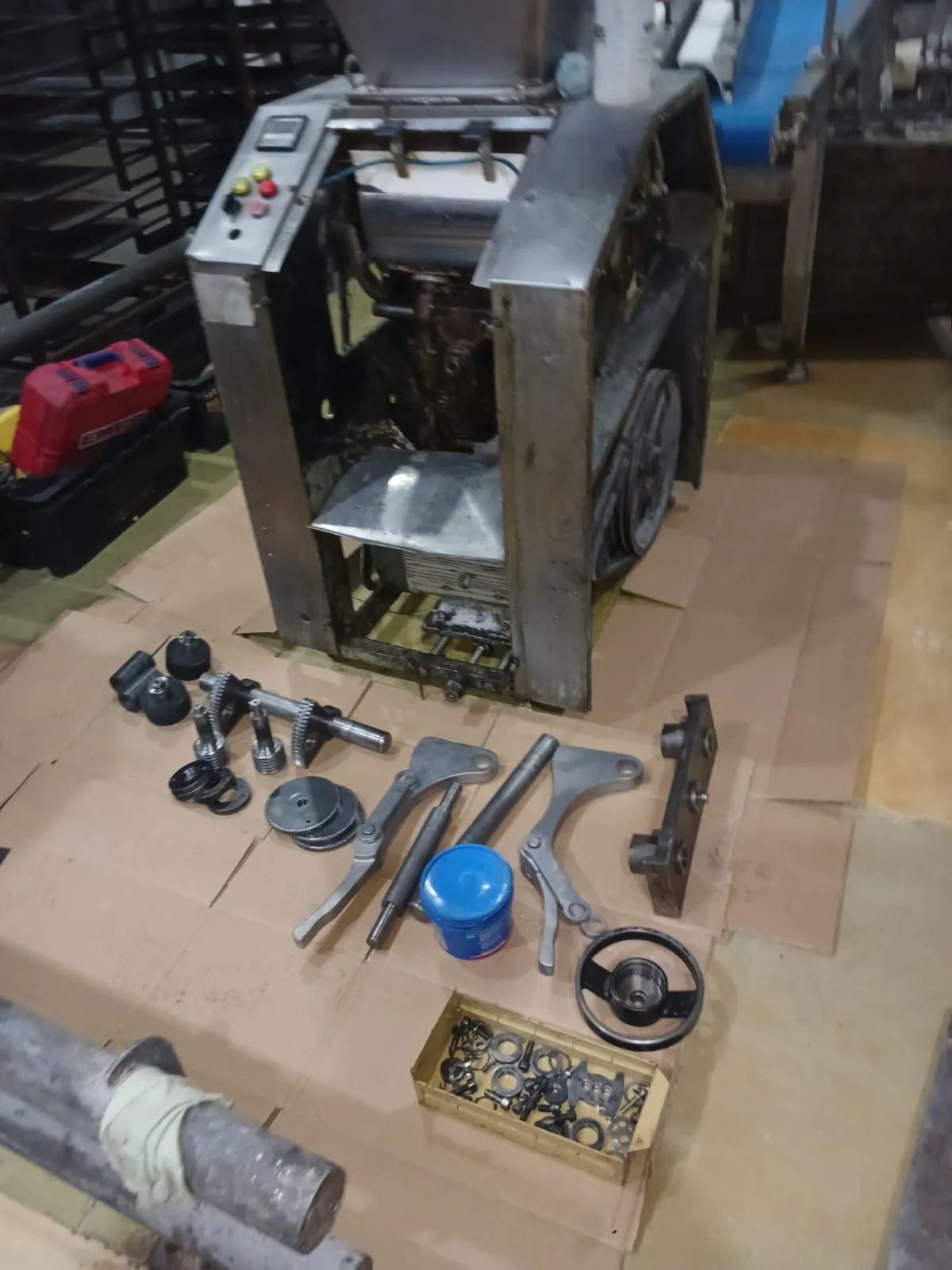 Piezas de maquinaria desmontadas, dispuestas sobre un suelo de baldosas junto a una gran máquina metálica.