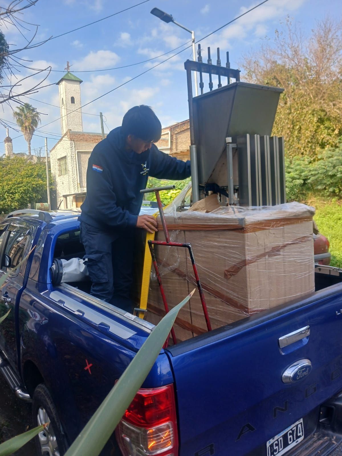 Un hombre carga una gran chimenea o estufa de metal en una camioneta azul al aire libre en un día soleado.