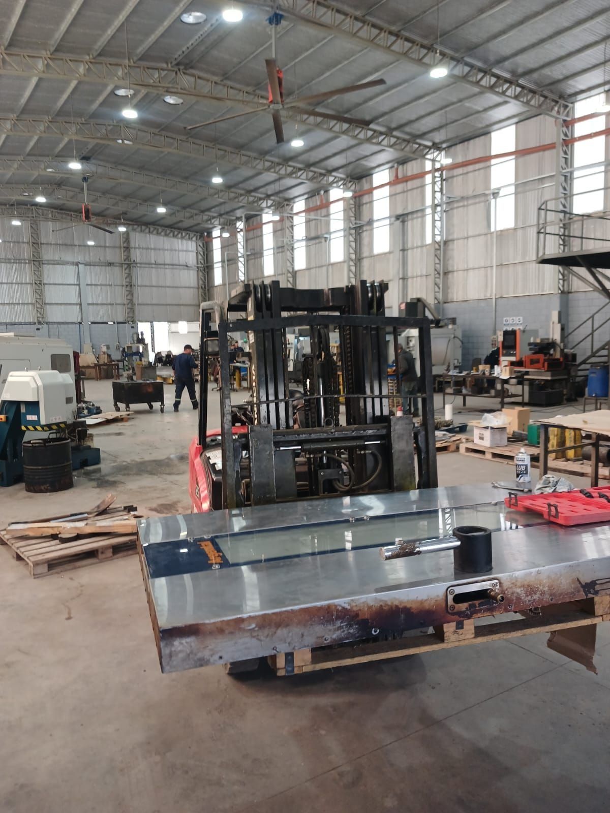Interior de un almacén con una carretilla elevadora que transporta un gran panel metálico cerca de palés y equipos apilados.