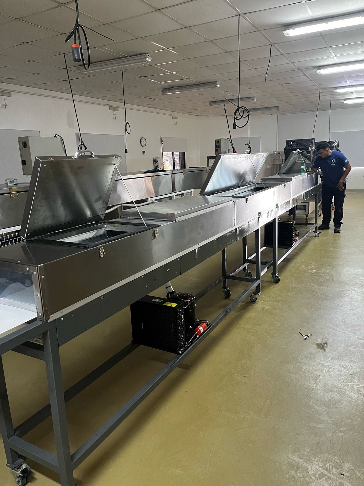Larga cinta transportadora metálica en una fábrica, con un operario de pie junto a ella bajo brillantes luces fluorescentes.