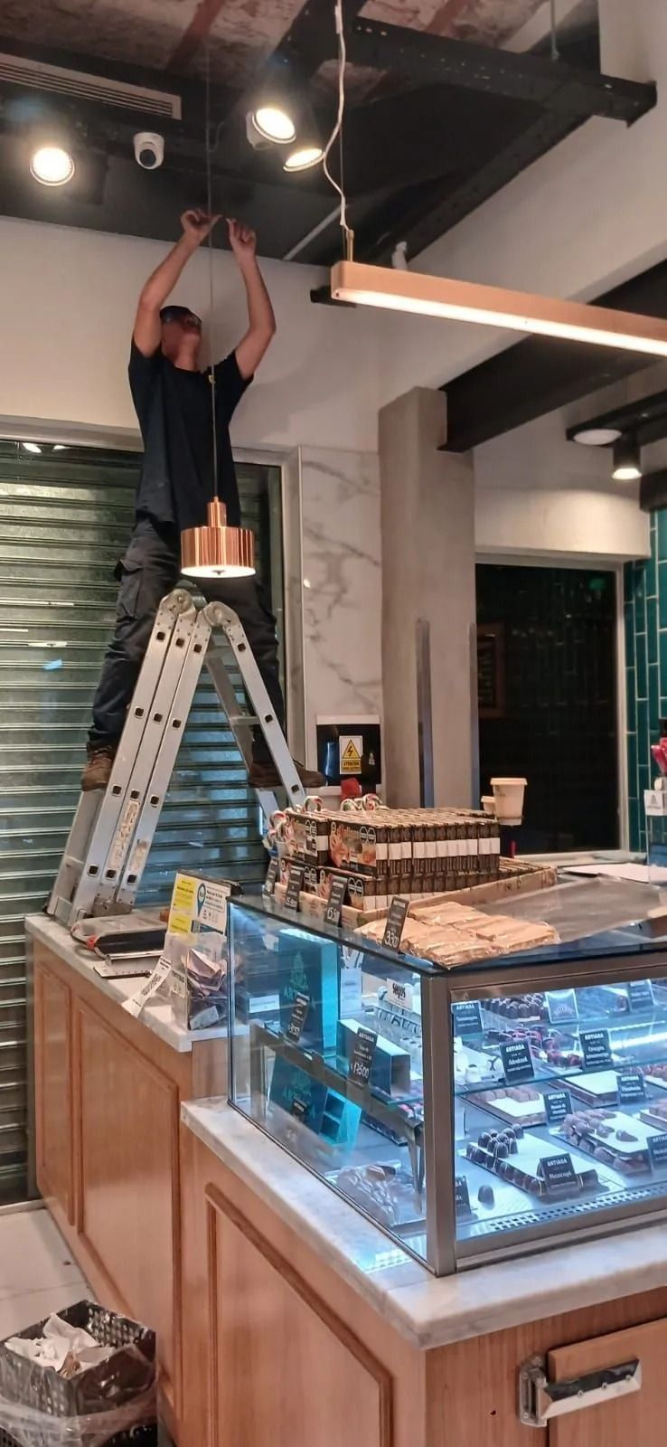 Un trabajador está subido a una escalera ajustando las luces del techo que hay encima de una vitrina de una panadería.