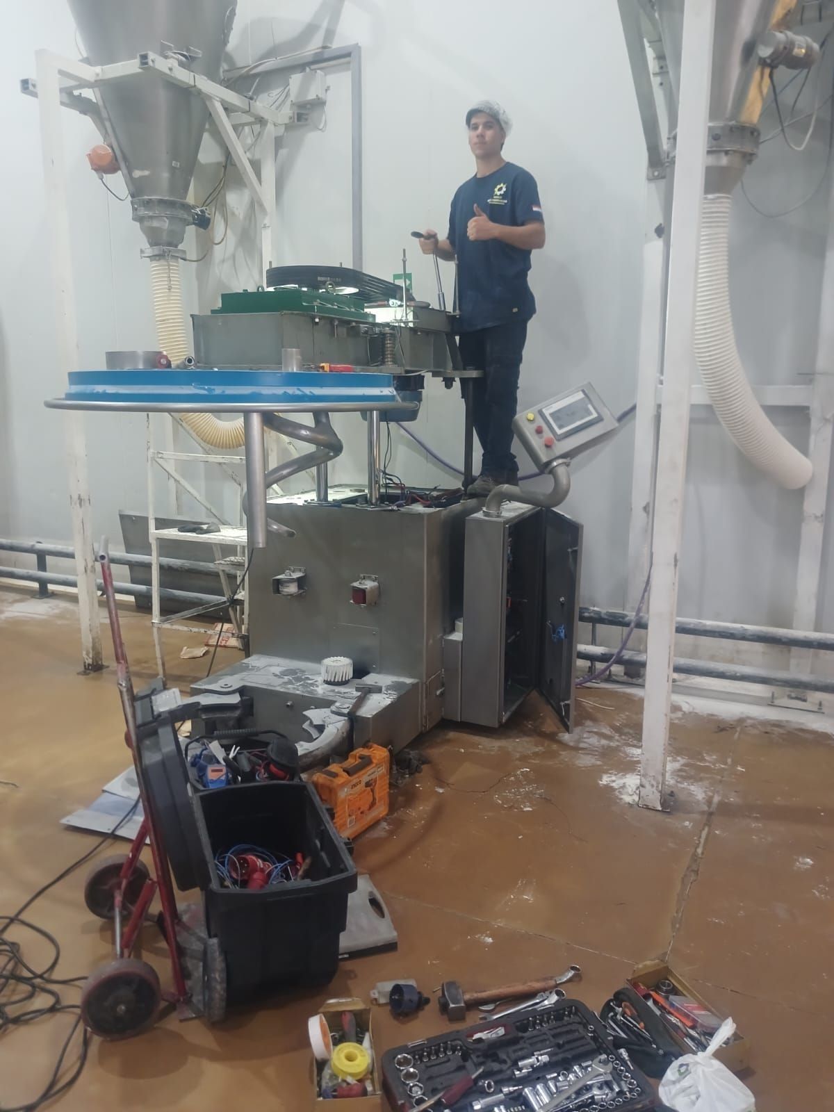 Persona de pie junto a maquinaria industrial en un taller, con herramientas y mangueras esparcidas por el suelo.