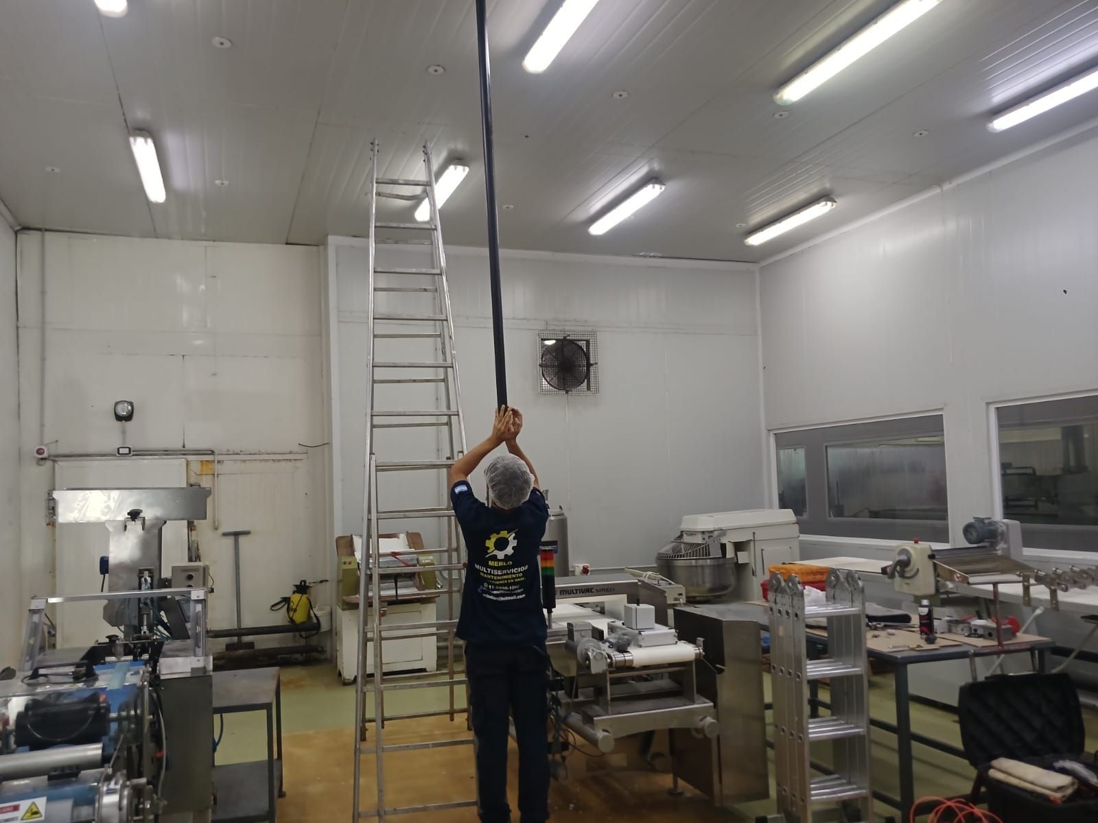 Una persona en un taller limpio se estira para alcanzar una escalera bajo luces brillantes del techo.