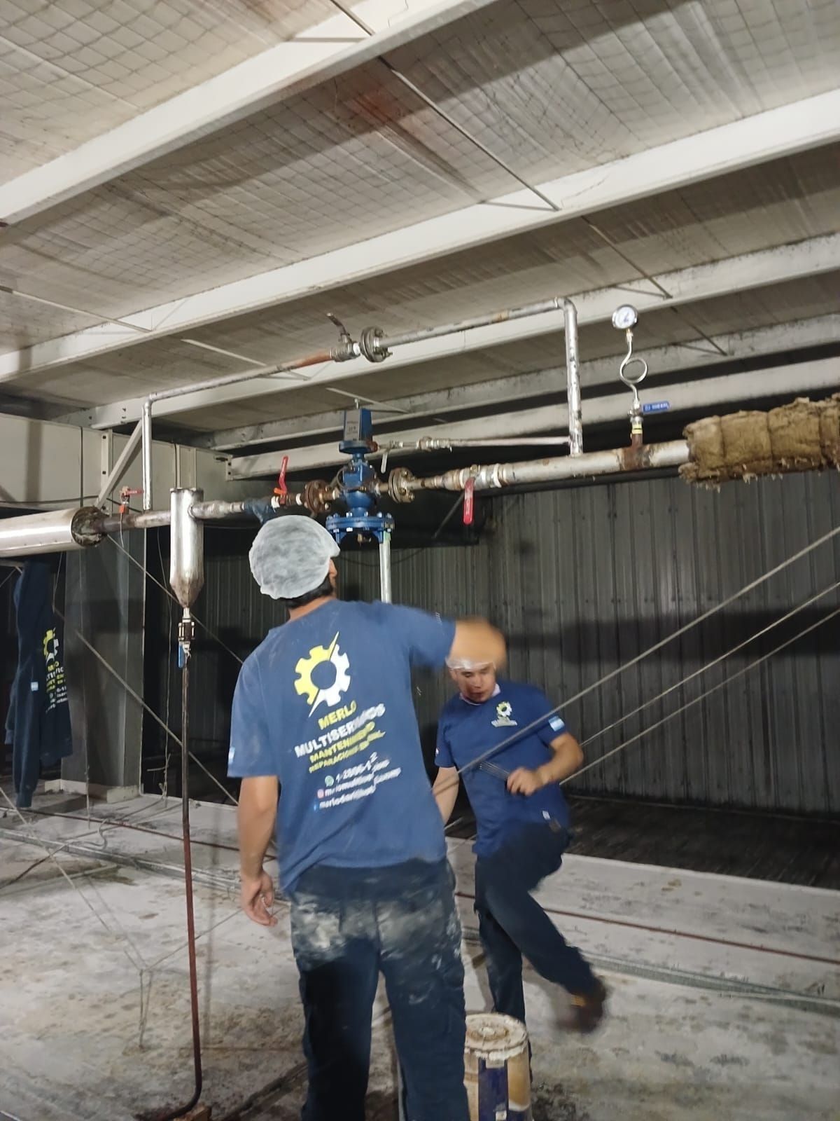 Dos trabajadores con camisas azules aplican pintura en aerosol dentro de un almacén industrial con tuberías en el techo.