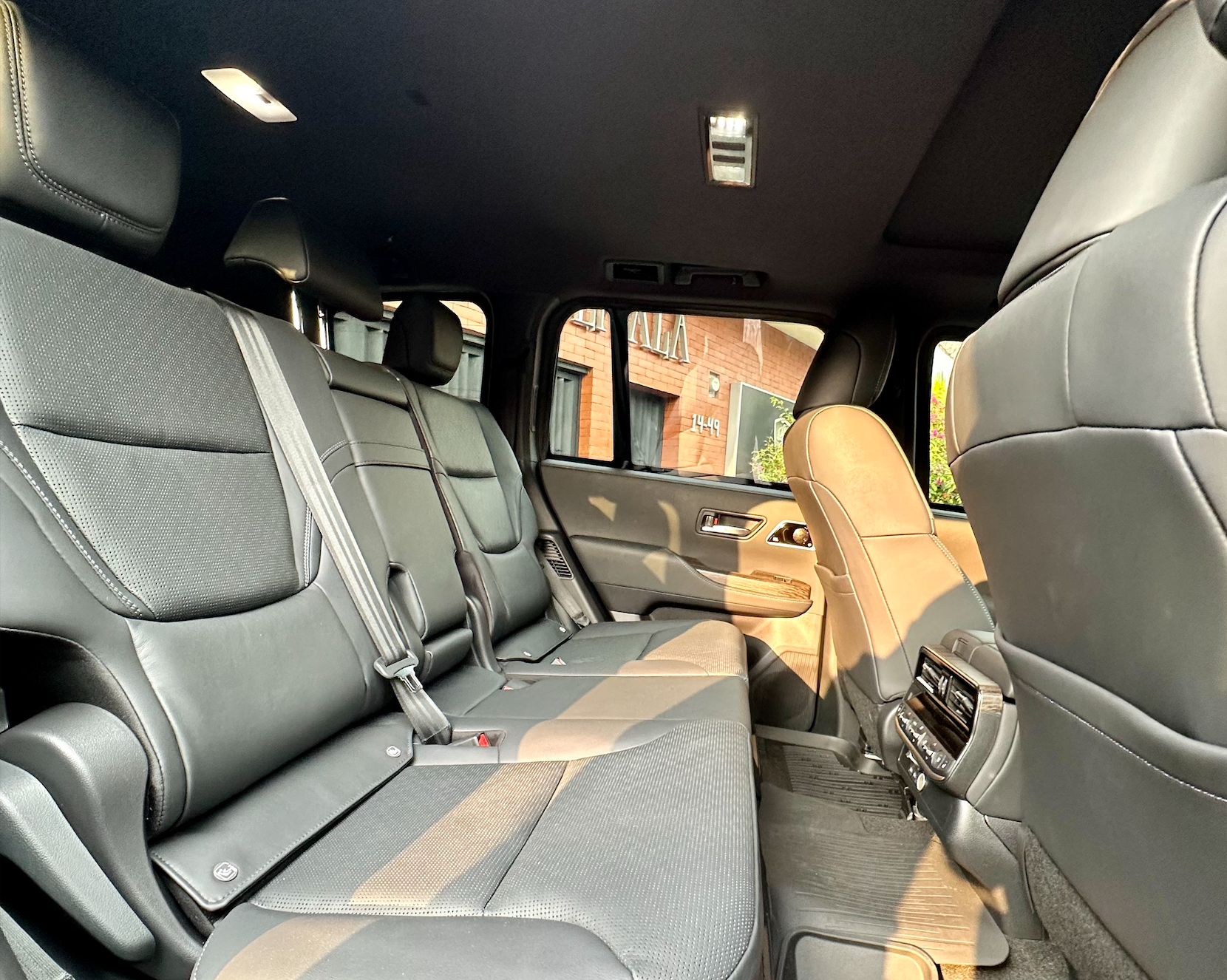 Interior de coche de cuero negro con tres asientos traseros y una puerta abierta a una calle soleada.