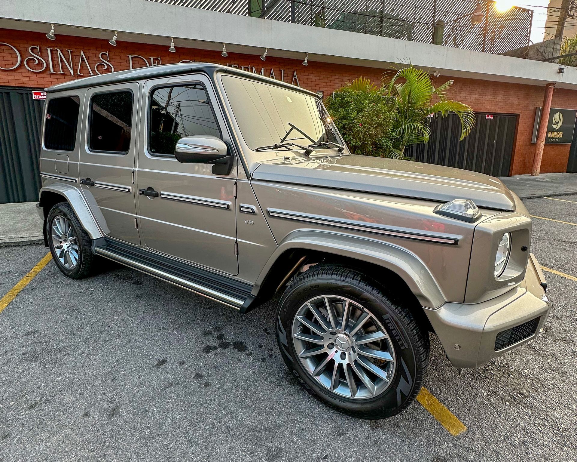Un SUV Mercedes-Benz Clase G de color beige estacionado en una calle frente a un edificio al atardecer.