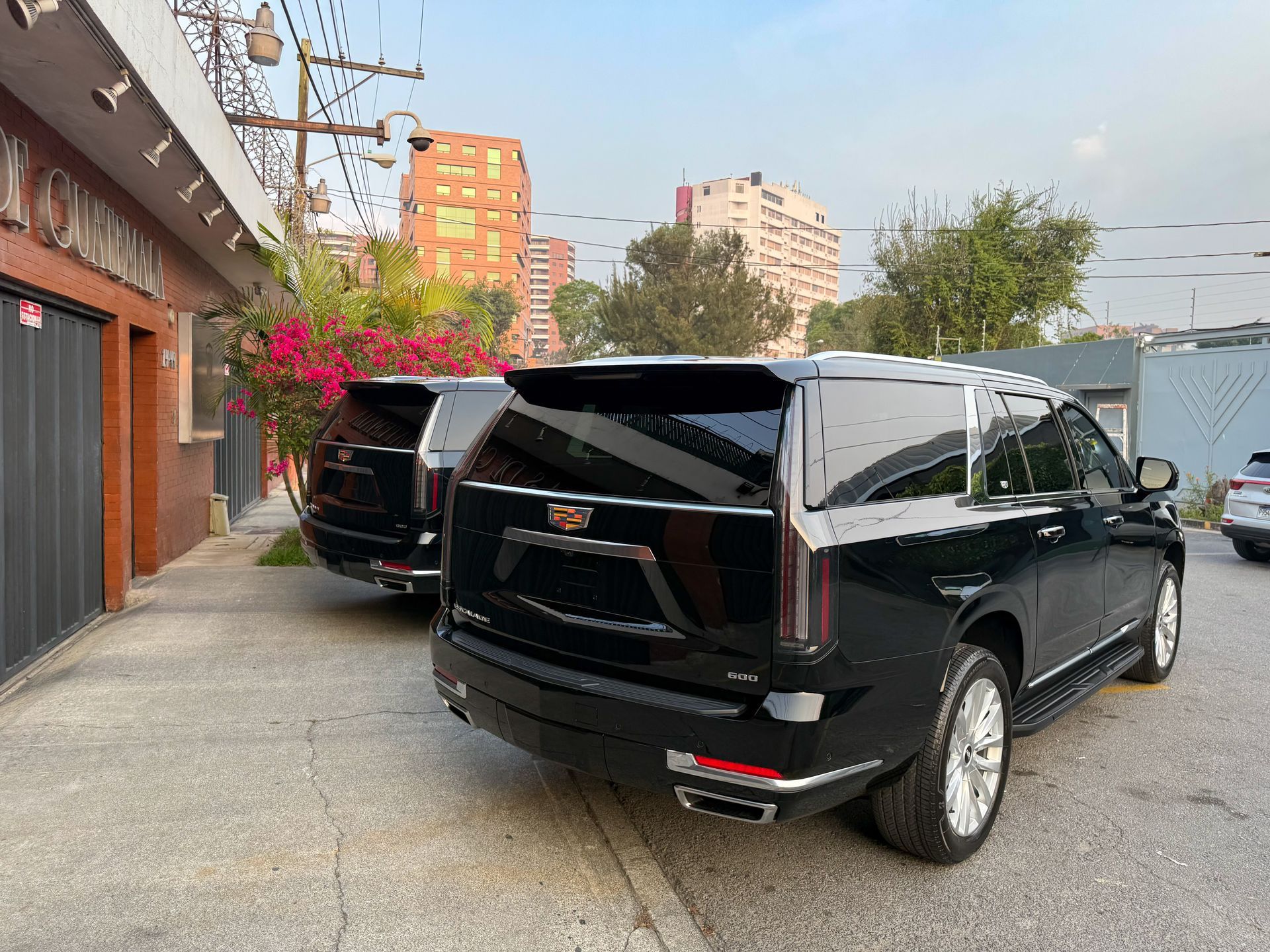 Dos camionetas SUV negras estacionadas junto a un edificio cerca de flores rosas colgantes en un estacionamiento pavimentado.