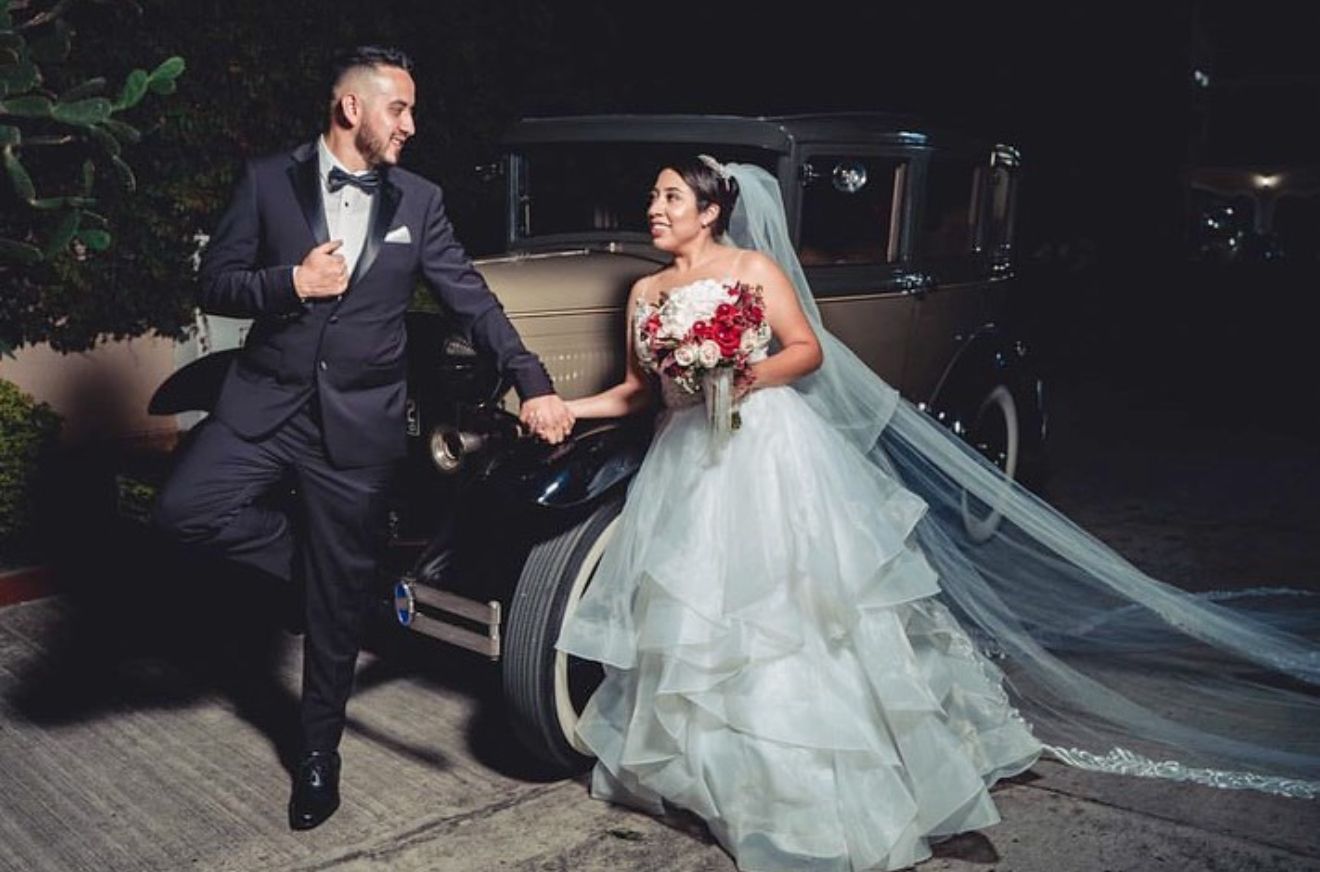 Novios tomados de la mano junto a un auto antiguo por la noche, sonriendo con sus trajes de boda.