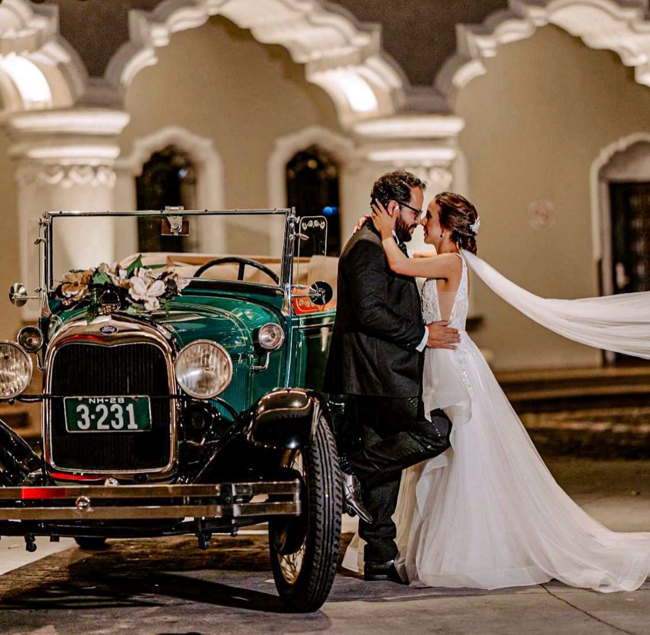 Novios abrazados junto a un coche verde antiguo en un patio ornamentado.