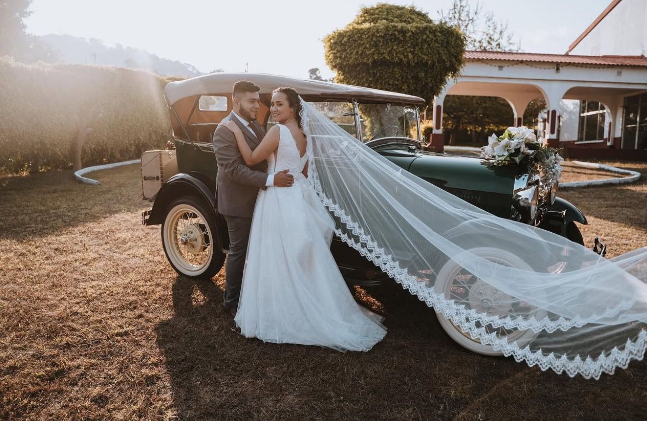Los novios se abrazan junto a un coche antiguo con un largo velo de encaje en un entorno exterior soleado.