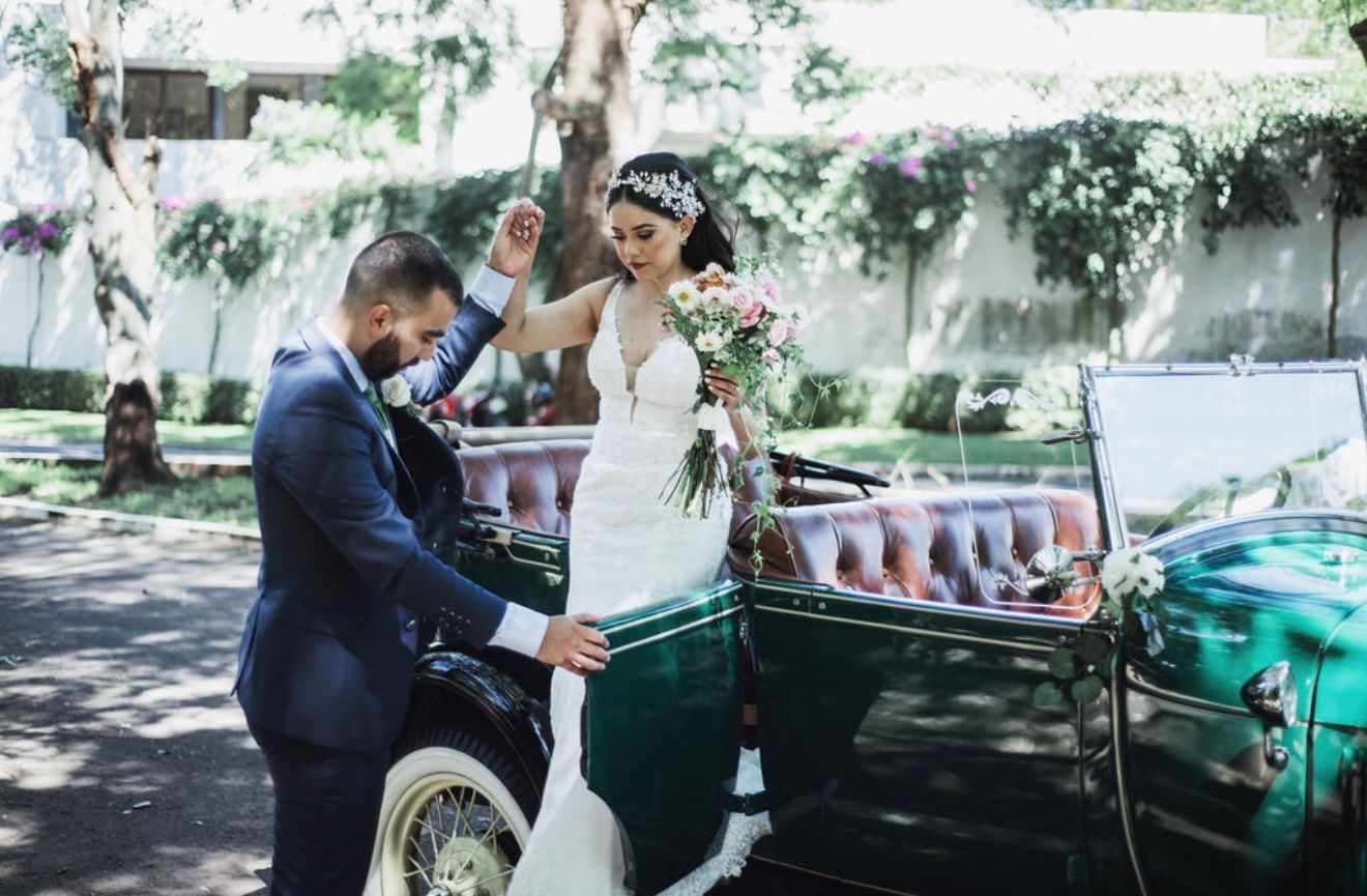 La novia, vestida de blanco, y el novio, con traje azul marino, bailan al aire libre junto a un coche antiguo