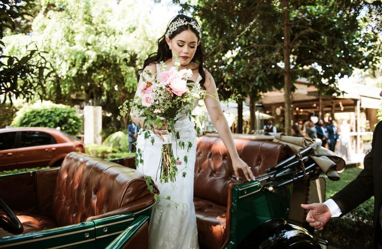 Una mujer con un vestido blanco estampado de flores sube a un coche verde antiguo al aire libre, sosteniendo un ramo.