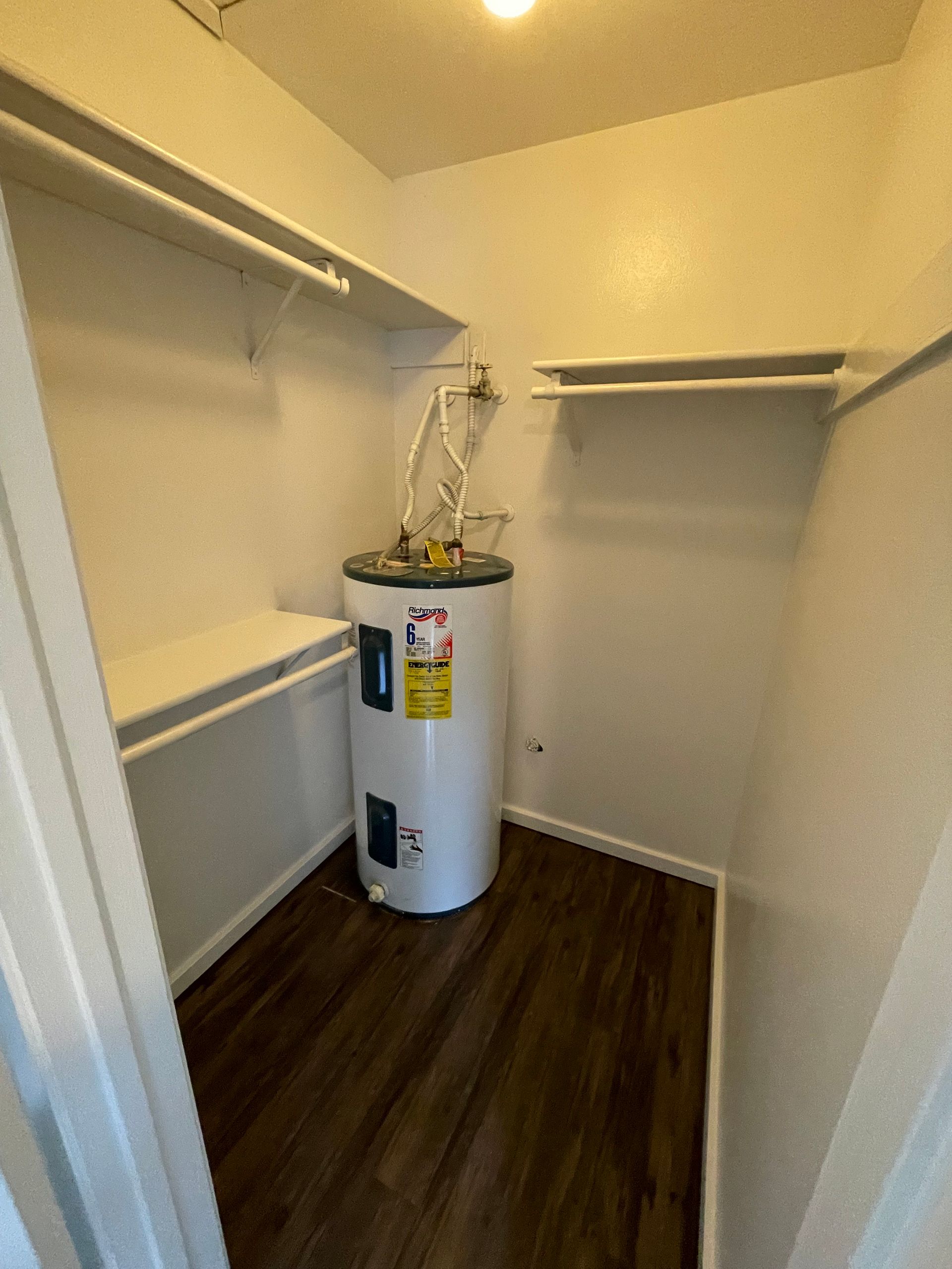 Small closet with a water heater, shelves, and a clothes rod. Dark wood-look floor, white walls.