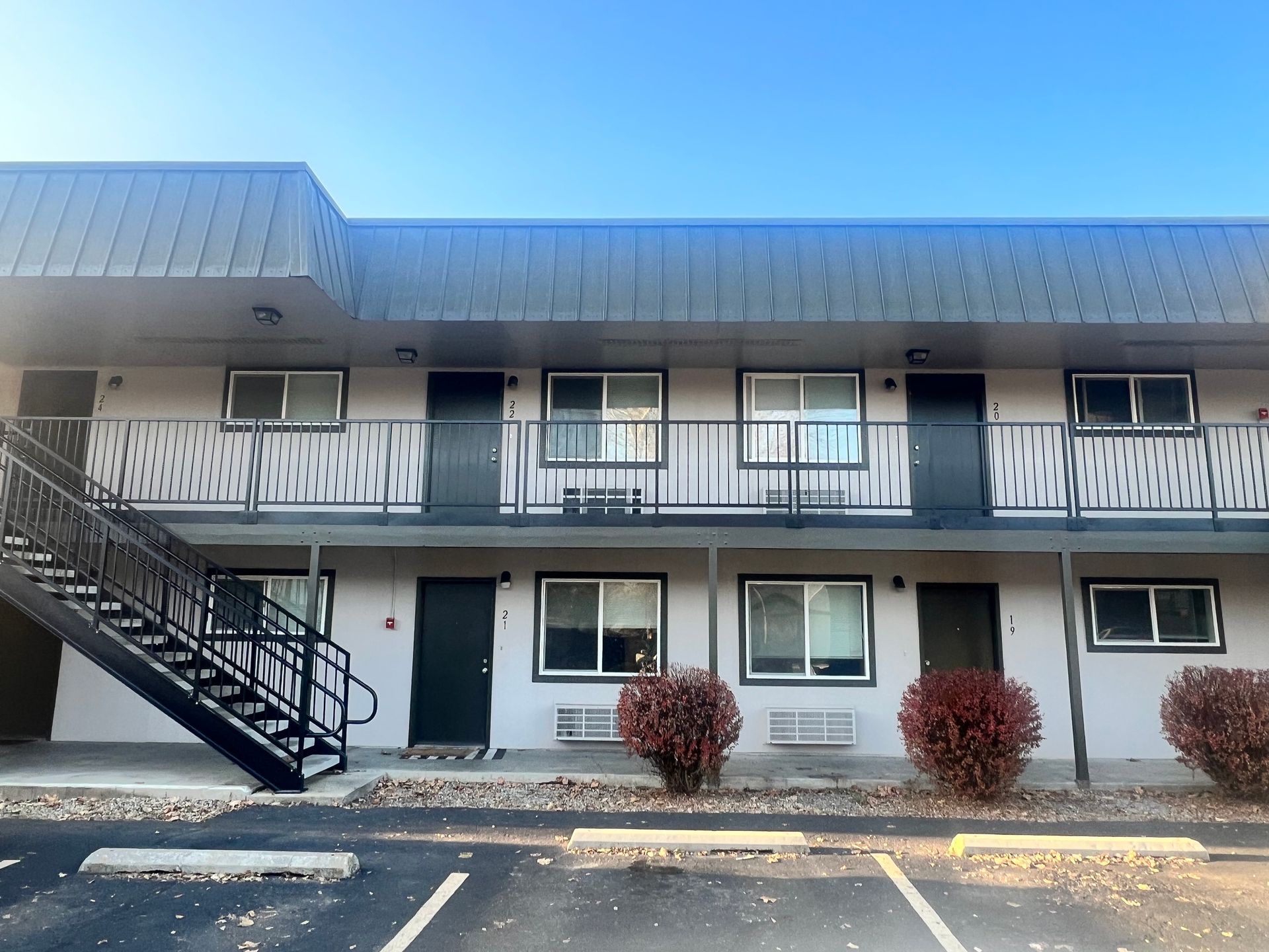 Two-story motel exterior with dark doors, metal stairwell, windows, and bushes against a blue sky.