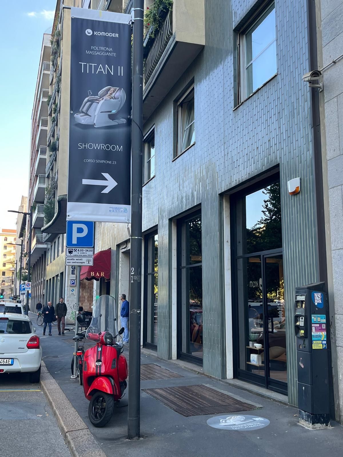 Via urbana con uno scooter rosso parcheggiato accanto a un edificio grigio e un cartello che indica l'ingresso di un negozio.