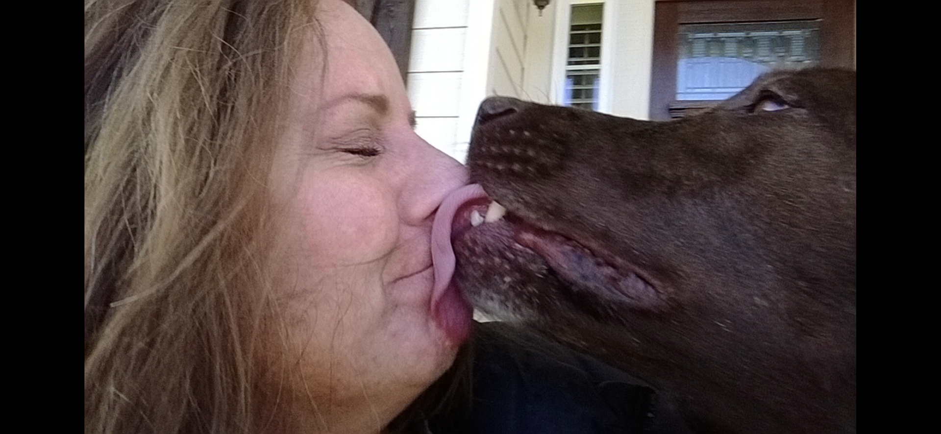 Woman being licked on the nose by a brown dog; both have closed eyes.