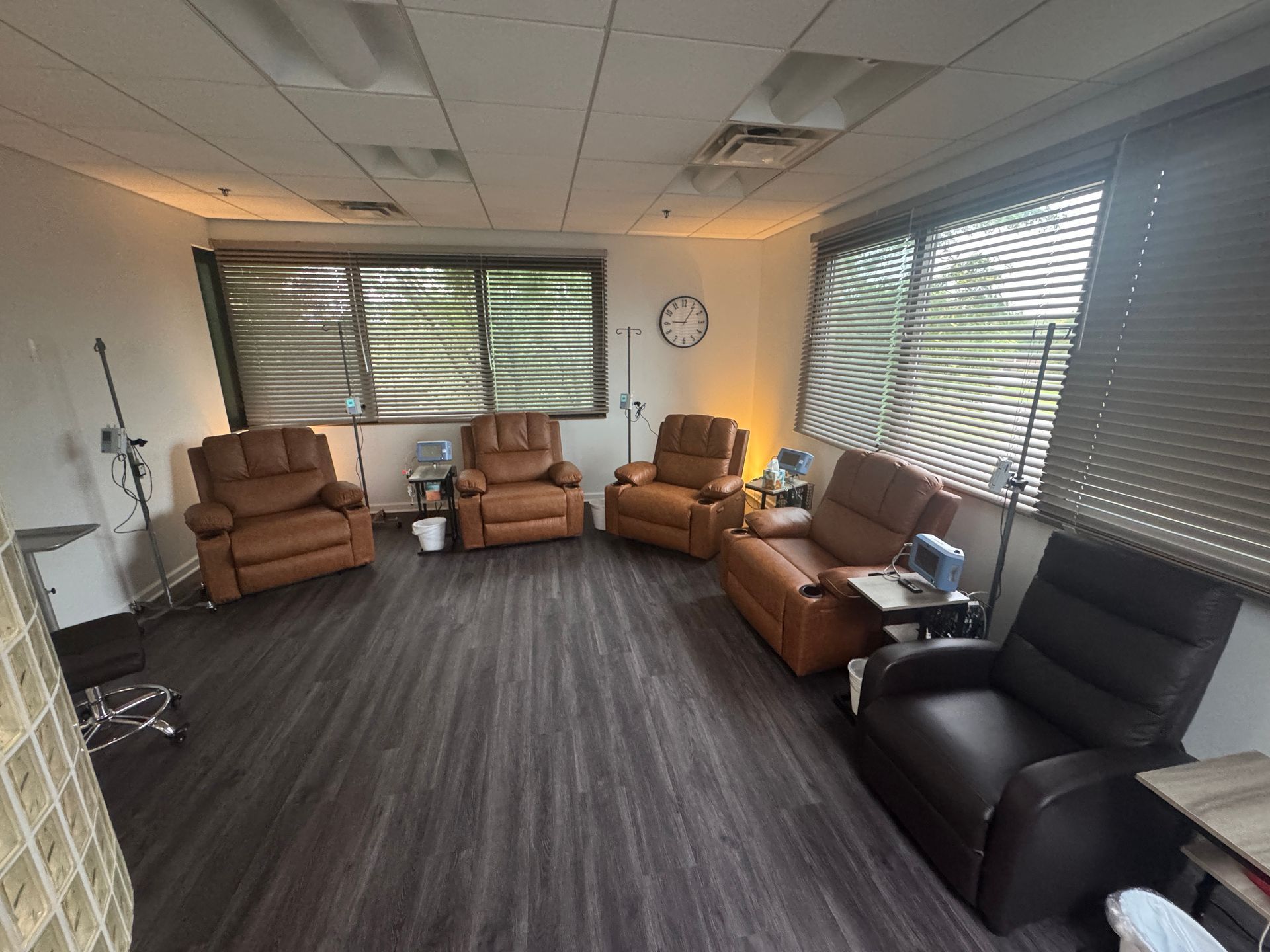 Empty lounge with brown and black recliners, gray carpet, large windows, and blinds