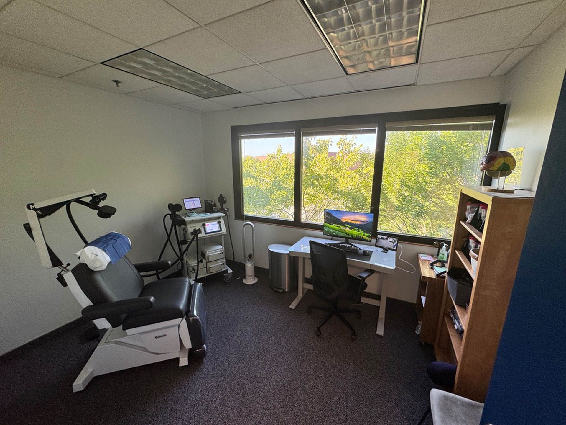 Medical office with a patient chair, TMS Therapy equipment, and a desk near a window overlooking trees.
