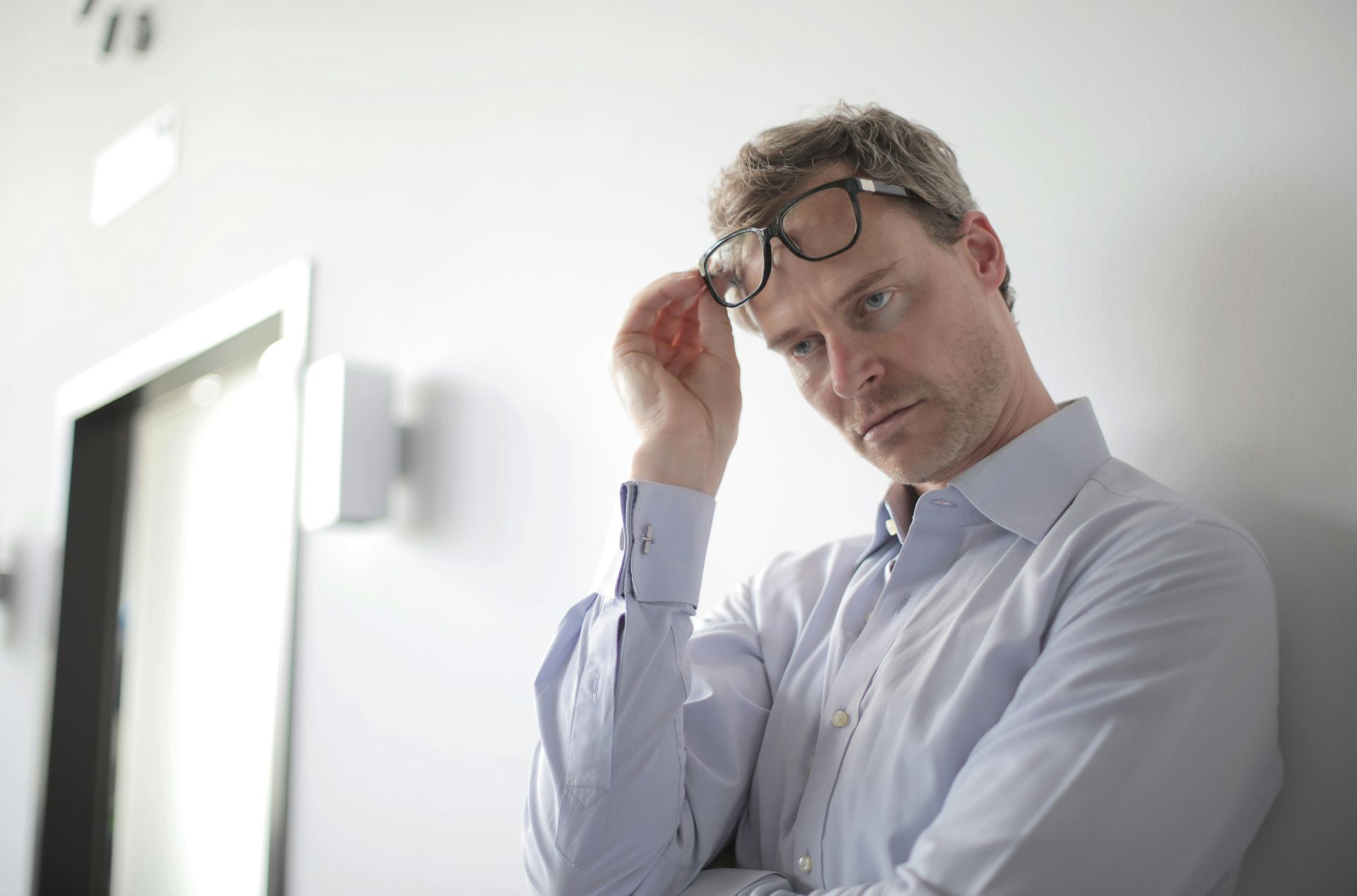 Man in a light blue shirt, holding glasses, leaning against a wall, looking down, appearing contemplative.
