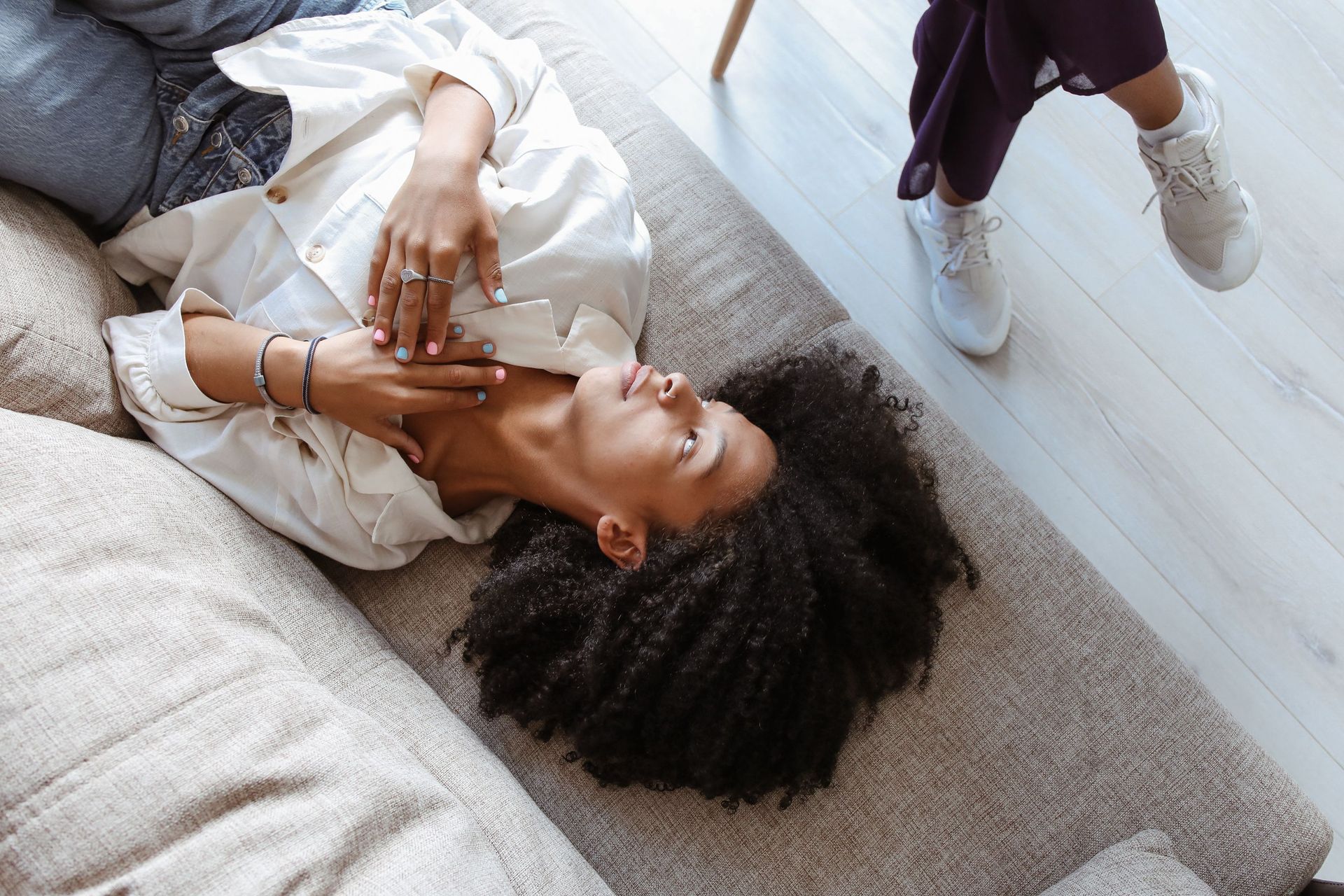 Person lying on a sofa with a hand over their face, beside a standing person in white sneakers on a light carpet.