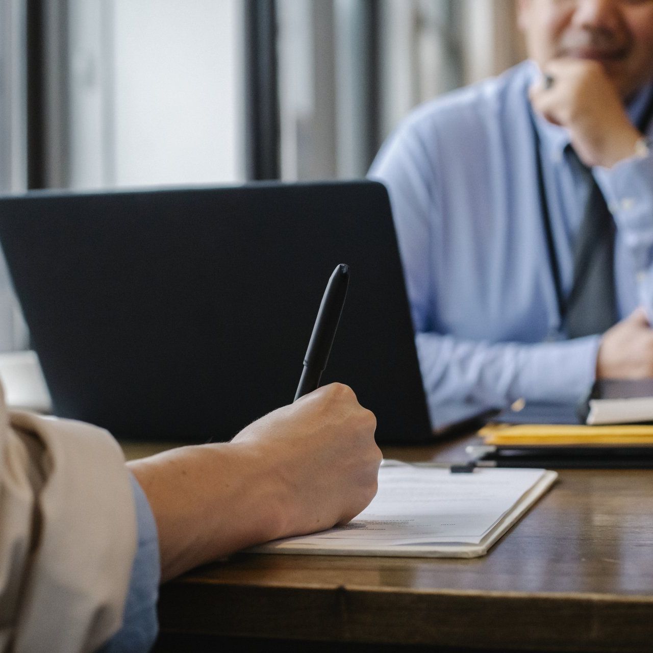 A person is writing on a piece of paper while another person looks at a laptop