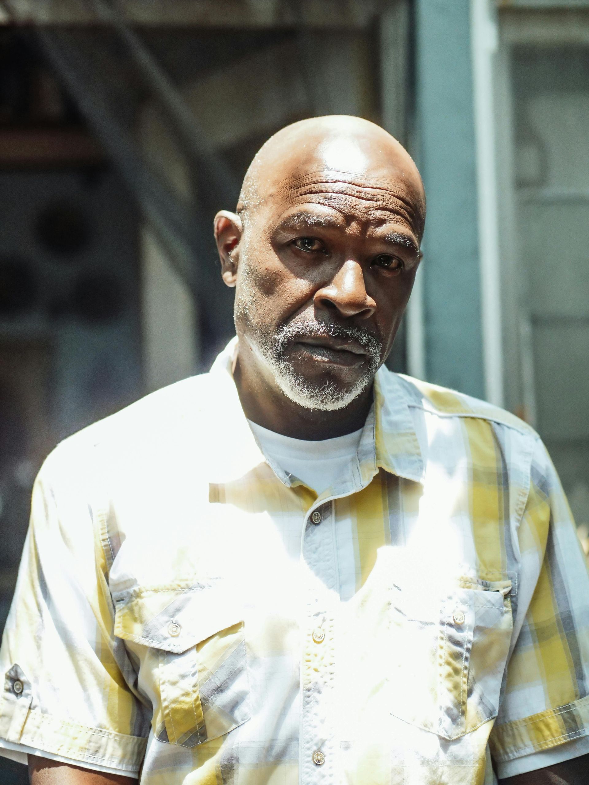 Bald Black man with gray beard, wearing yellow plaid shirt, looking at the camera, outdoors.