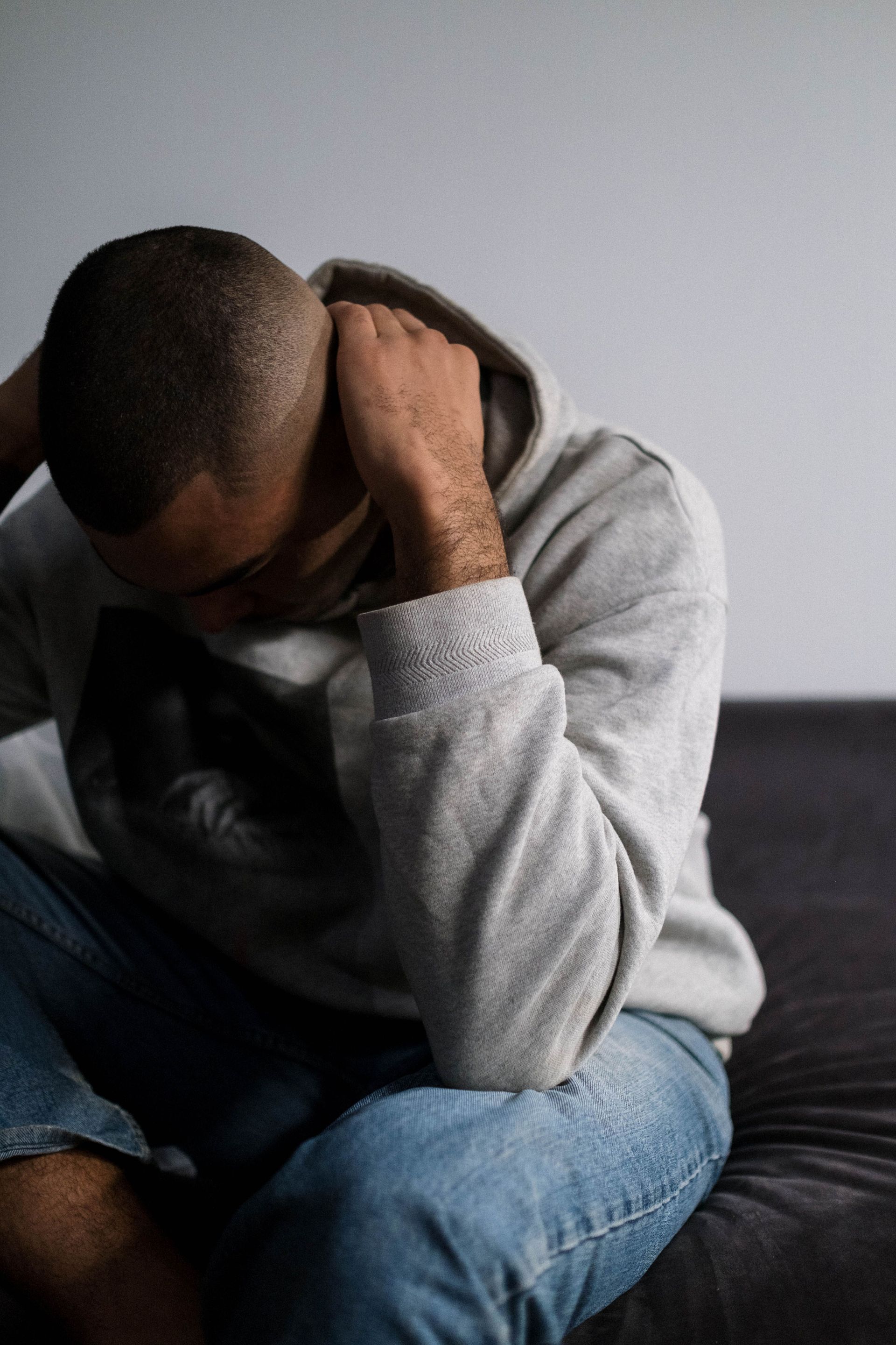 Person sitting on a bed, head bowed and hands covering their face, wearing a gray hoodie and jeans.