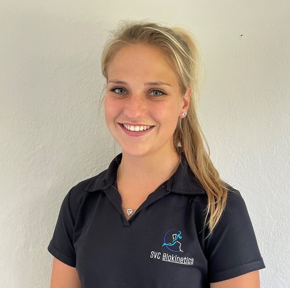 Woman with blonde hair smiling, wearing a black polo shirt with a logo, against a white wall.
