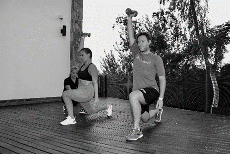Two people doing lunges with dumbbells on a wooden deck. One person raises a dumbbell overhead.