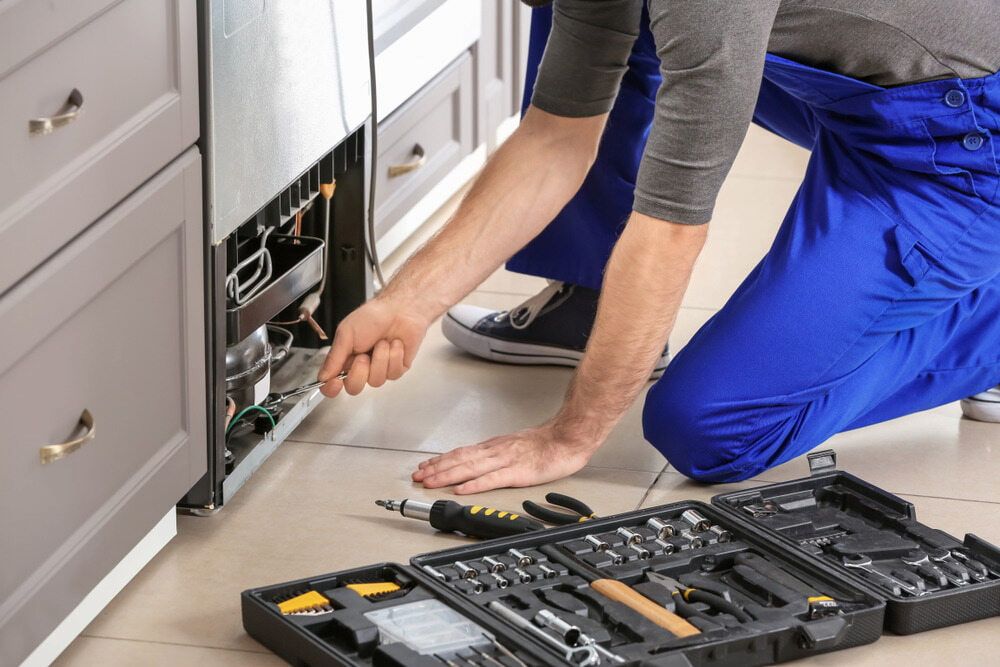 A Man is Kneeling on the Floor Fixing a Refrigerator — Tr!pl3 Fire Electrical & Contracting in Alice Springs, NT