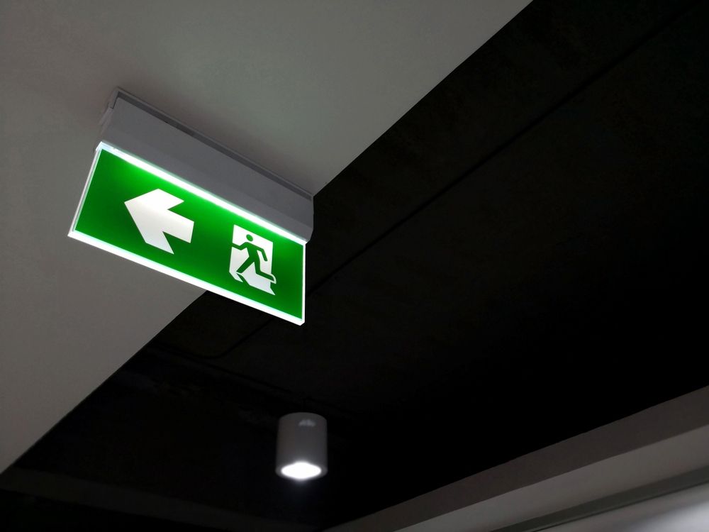 A Green Exit Sign is Hanging From the Ceiling in a Dark Room — Tr!pl3 Fire Electrical & Contracting in Alice Springs, NT
