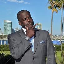 Man in gray suit pointing finger up, looking thoughtful outdoors near buildings and palms.
