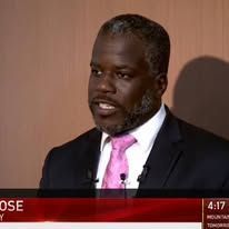 Man in a dark suit speaks, wearing a pink tie, in front of a wooden backdrop.