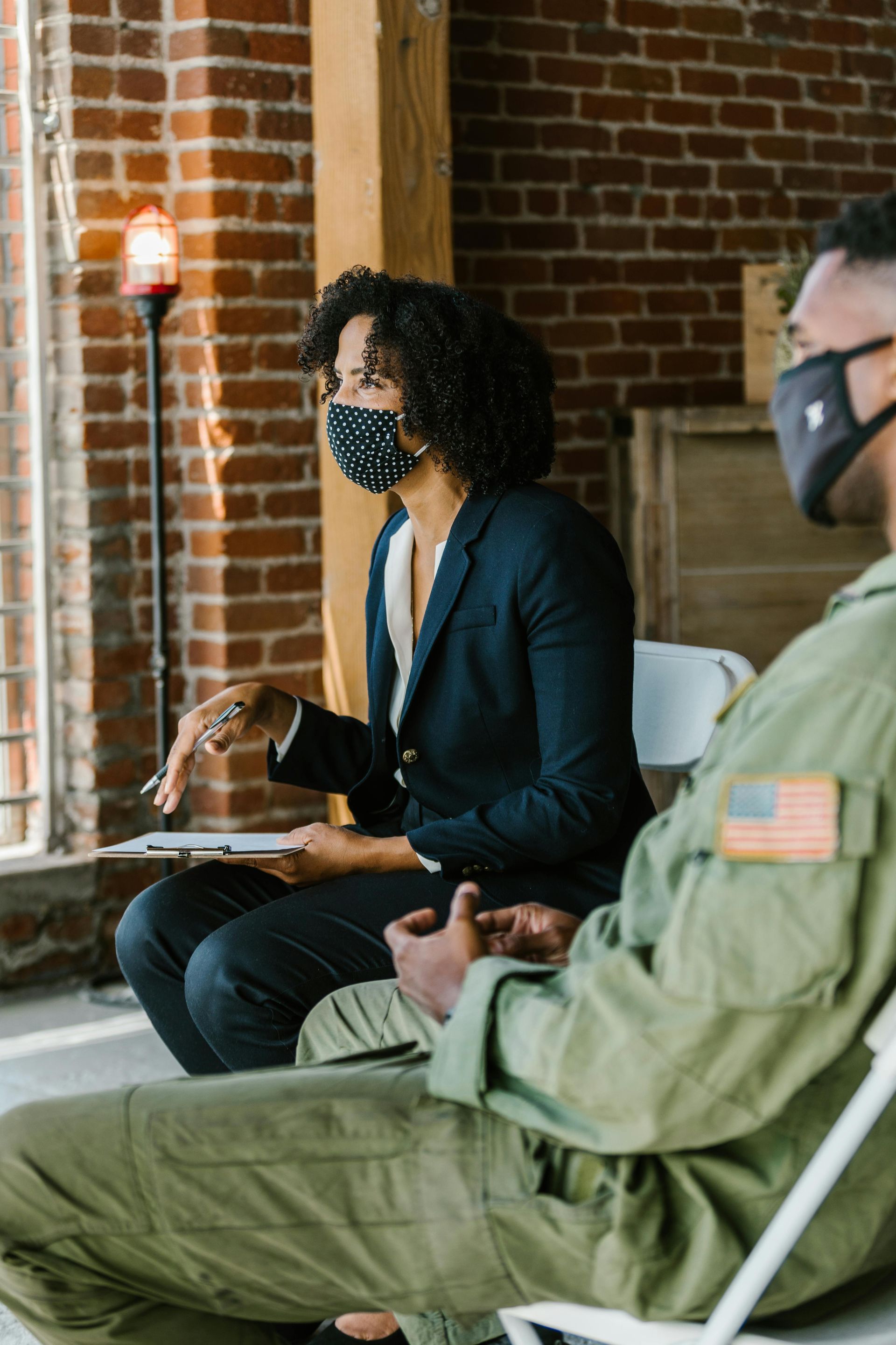 Woman in blazer and mask talking to person in military uniform, both seated indoors.