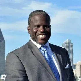 Man in a suit smiles in front of a city skyline, wearing a blue tie and pocket square.