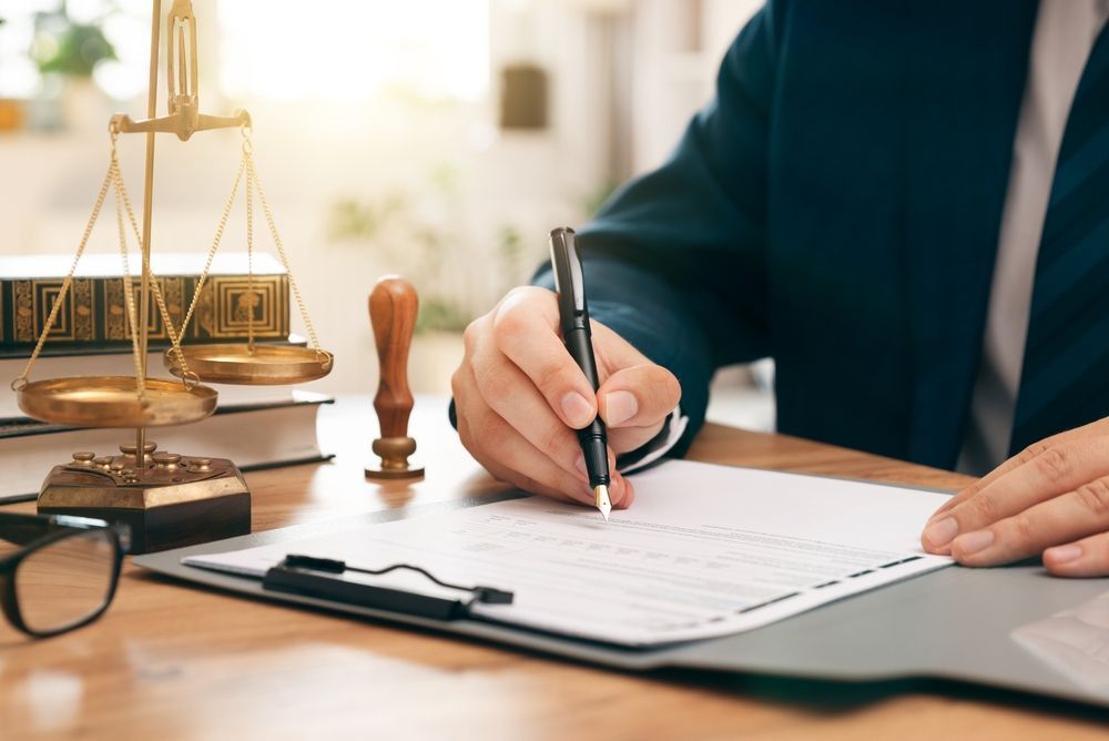 Person in a blue suit signs a document, scales of justice and law books on the desk.