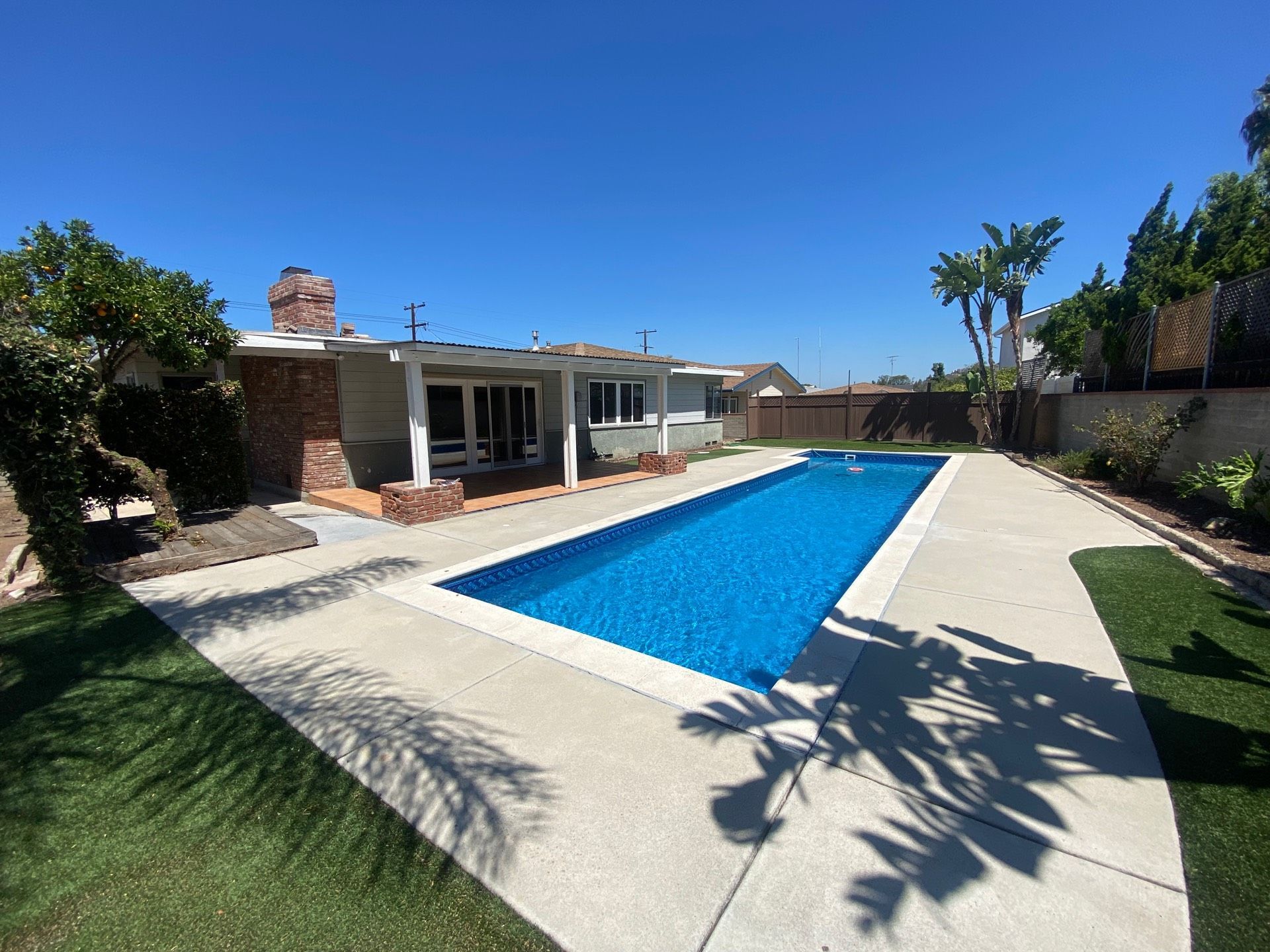 Backyard with long blue pool, concrete patio, lawn, and single-story house under a bright blue sky.