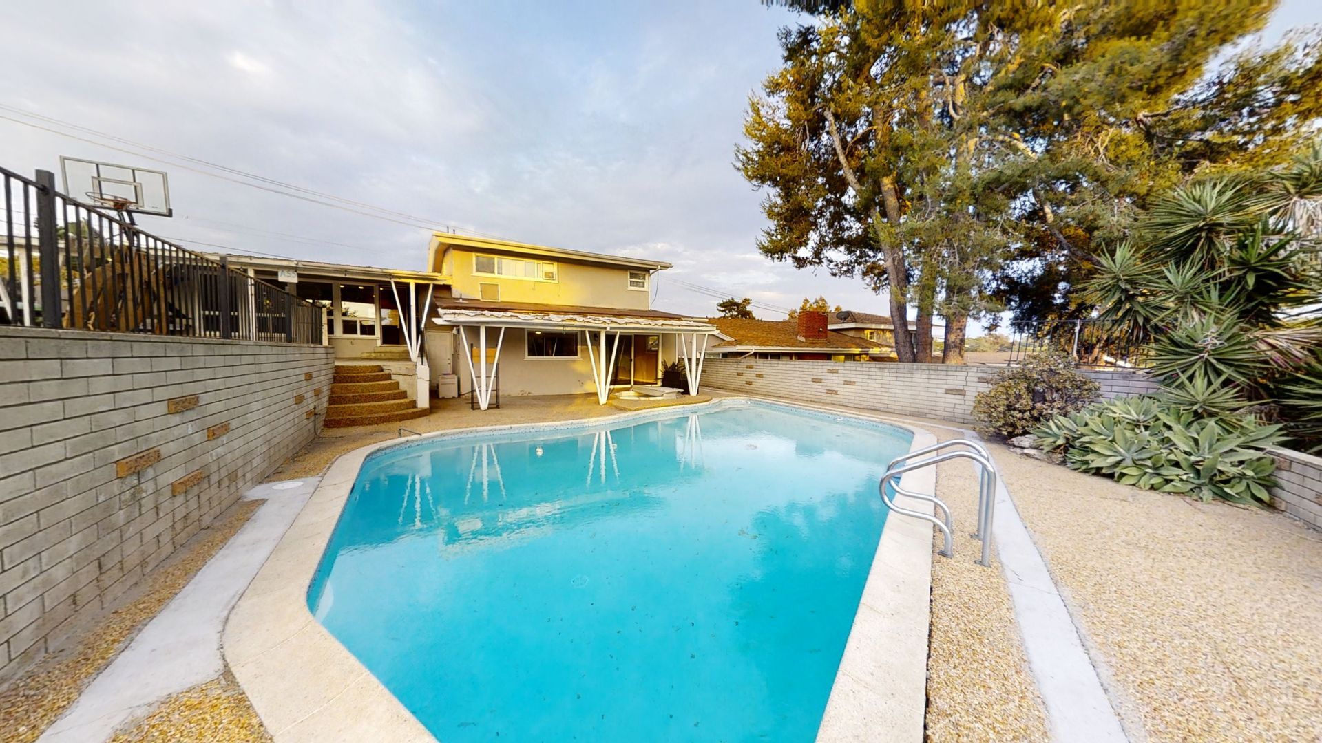 Backyard with a swimming pool, house, basketball hoop, and trees under a cloudy sky.