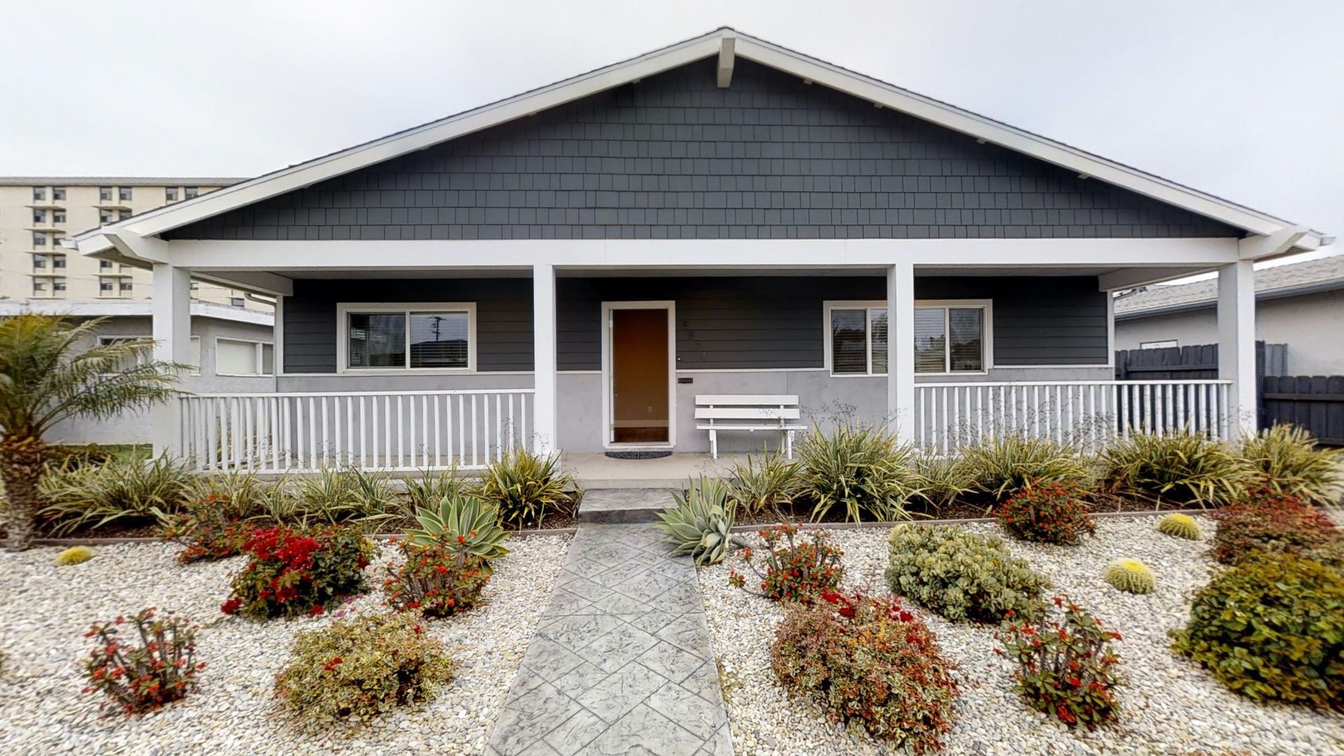 Gray house with porch, white railings, and gravel path in front of colorful bushes.