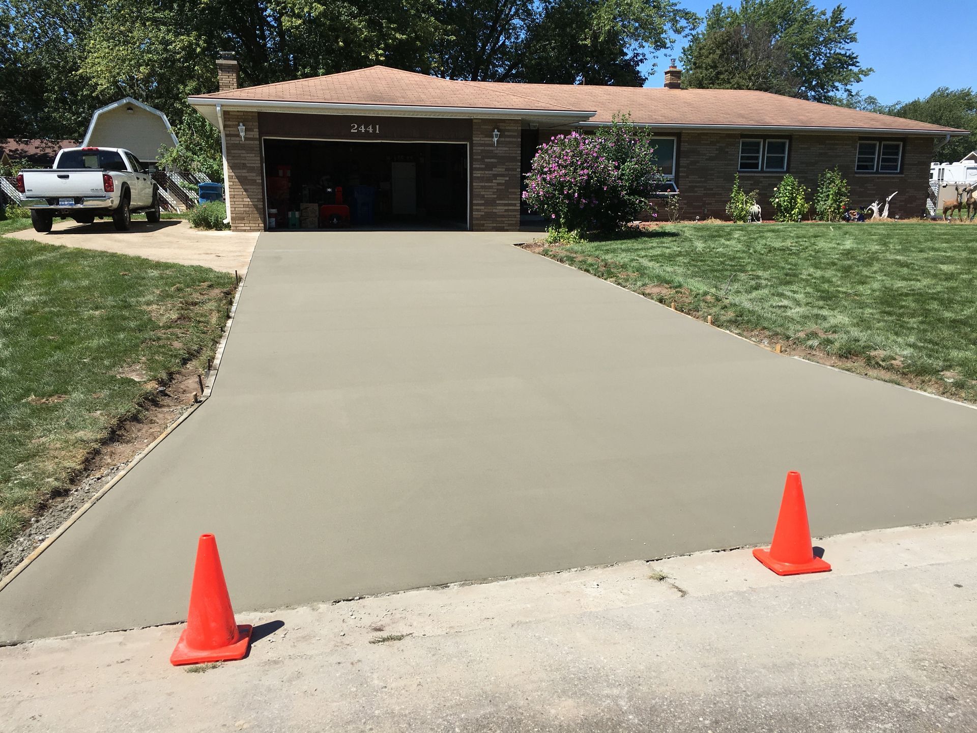 Newly poured concrete driveway in front of a brick house, with orange safety cones on the street.