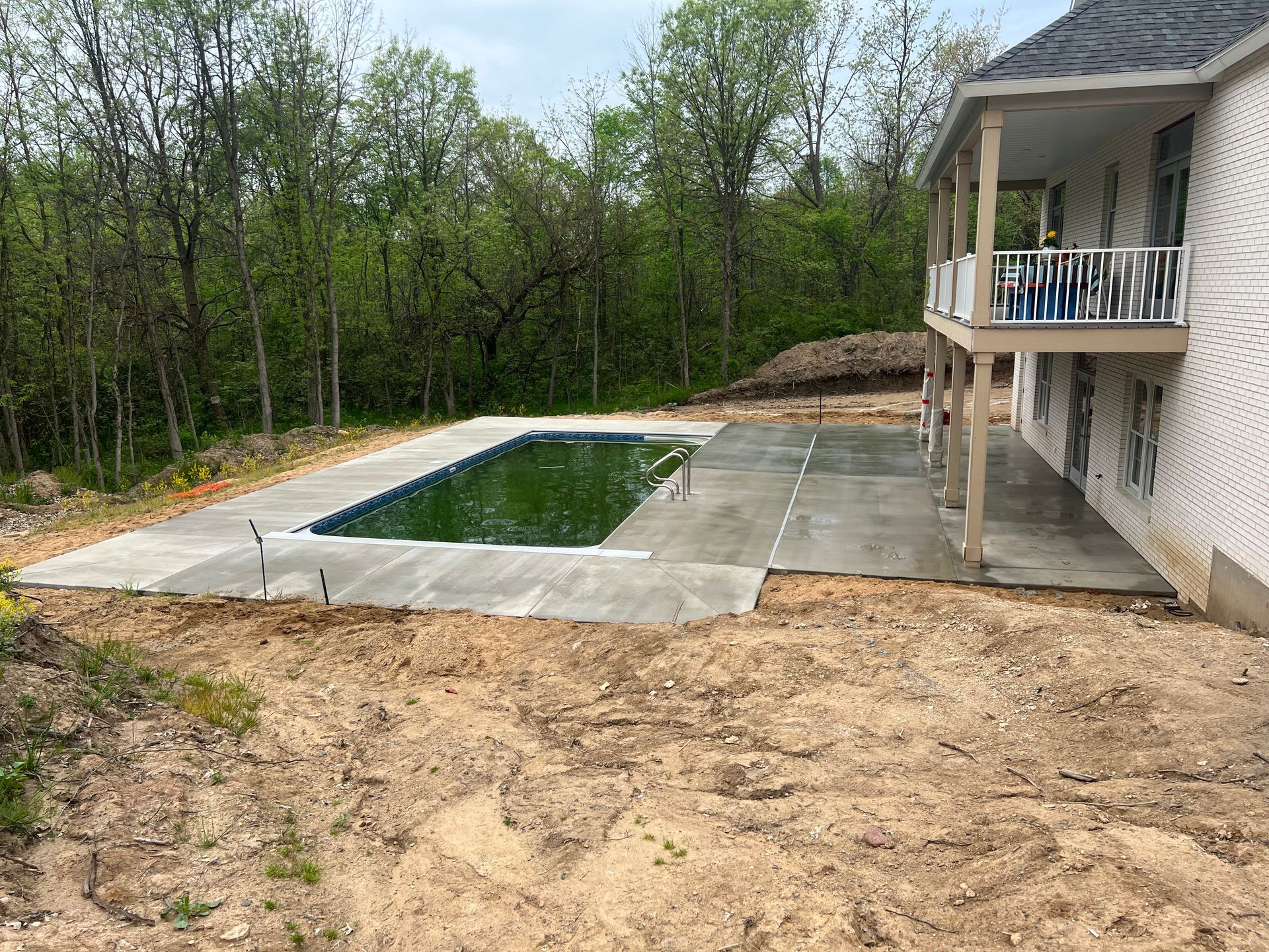 Concrete patio with unfinished swimming pool next to a house with a balcony and surrounding trees.