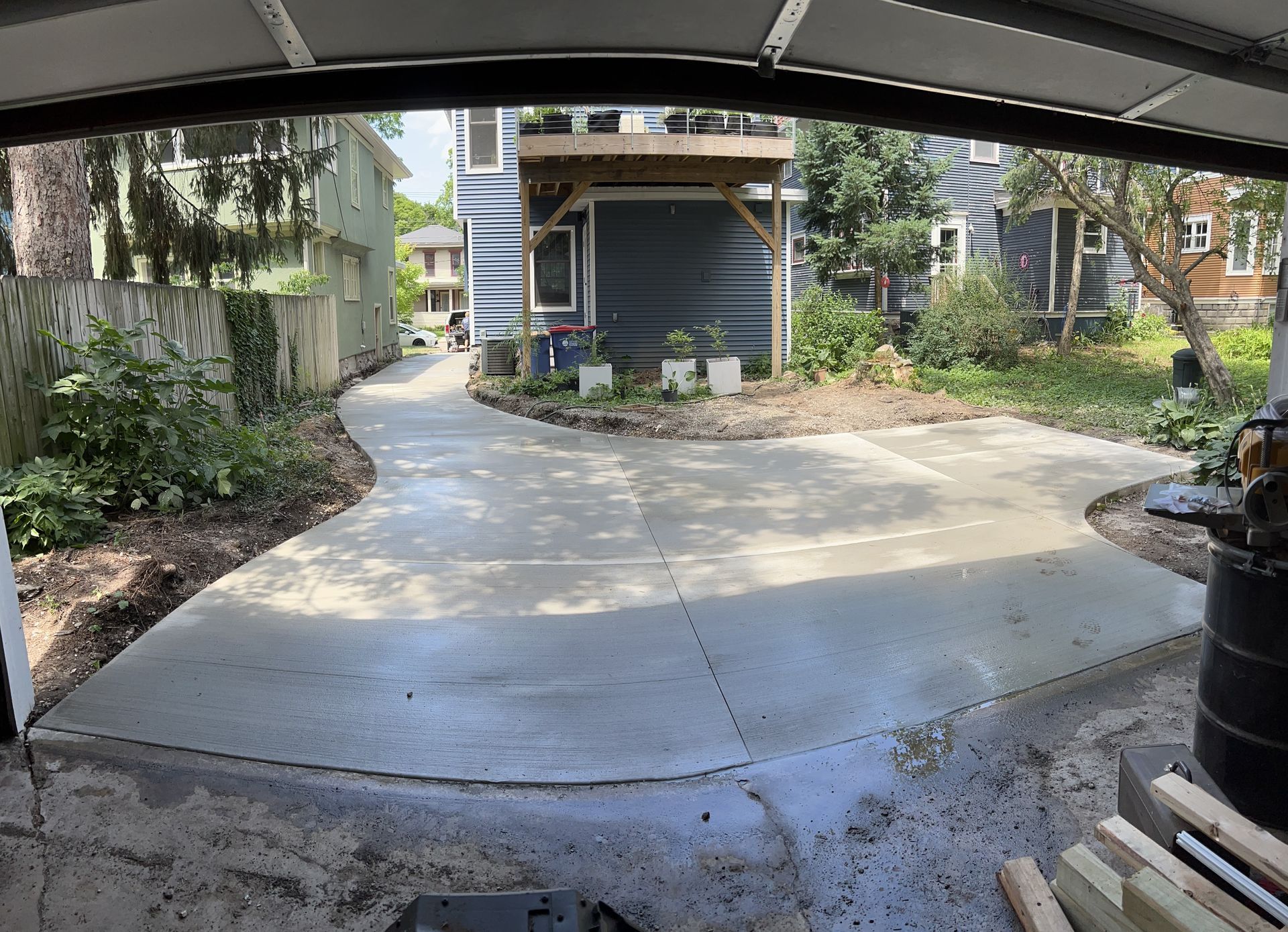 Concrete driveway curves towards a house with a deck; trees and green foliage line the sides.