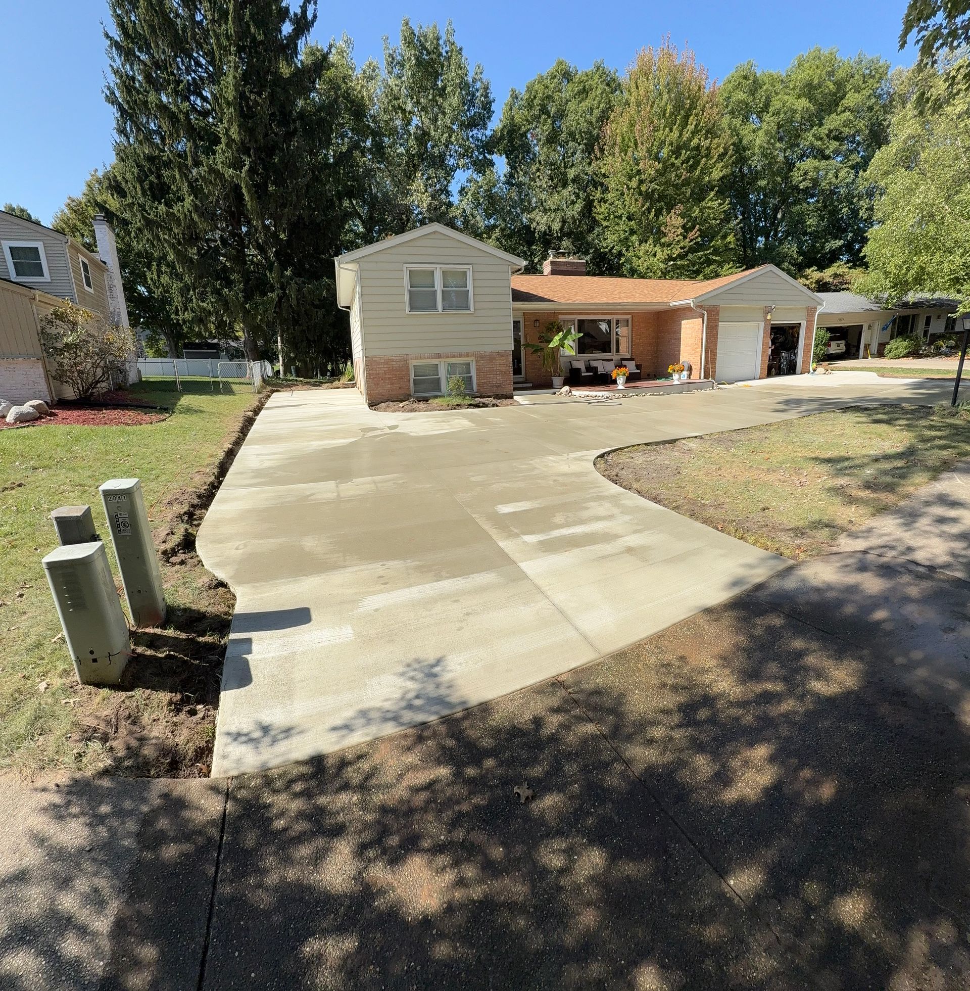 A newly poured concrete driveway in front of a tan brick and beige house with a garage and green trees.