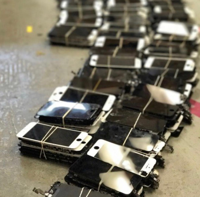 Stacks of black smartphones with shattered screens arranged on a floor in a warehouse-like setting