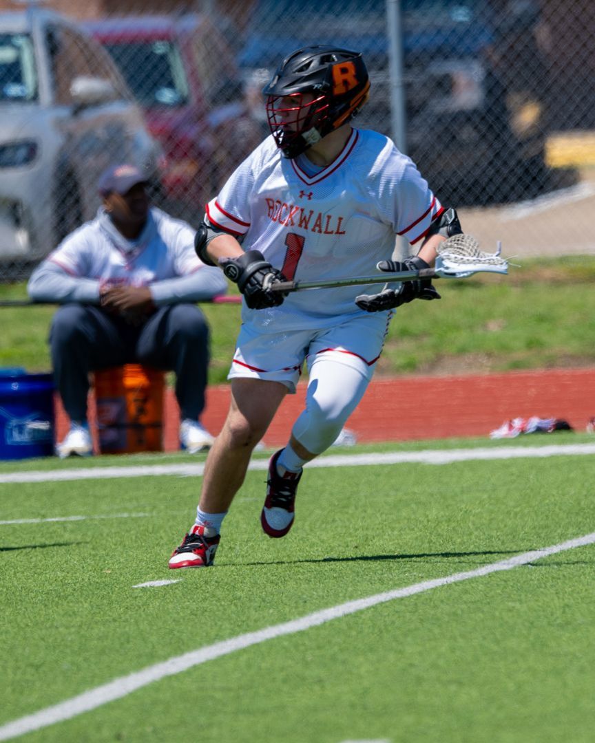 Lacrosse player in white and orange uniform running on a turf field with a stick, sideline in background